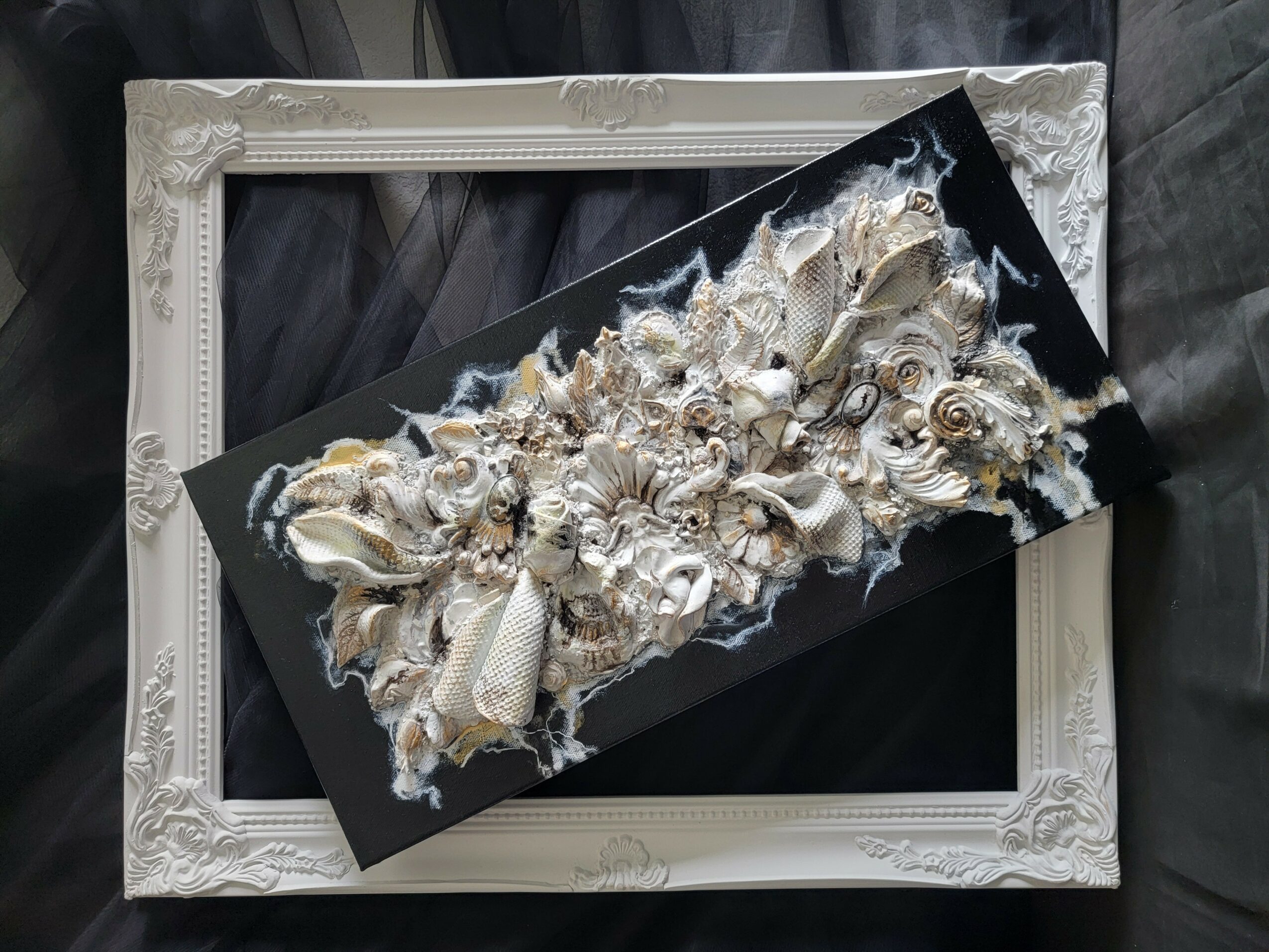 Decorative black and white floral artwork on a rectangular canvas, placed on a white ornate frame, on a dark surface.