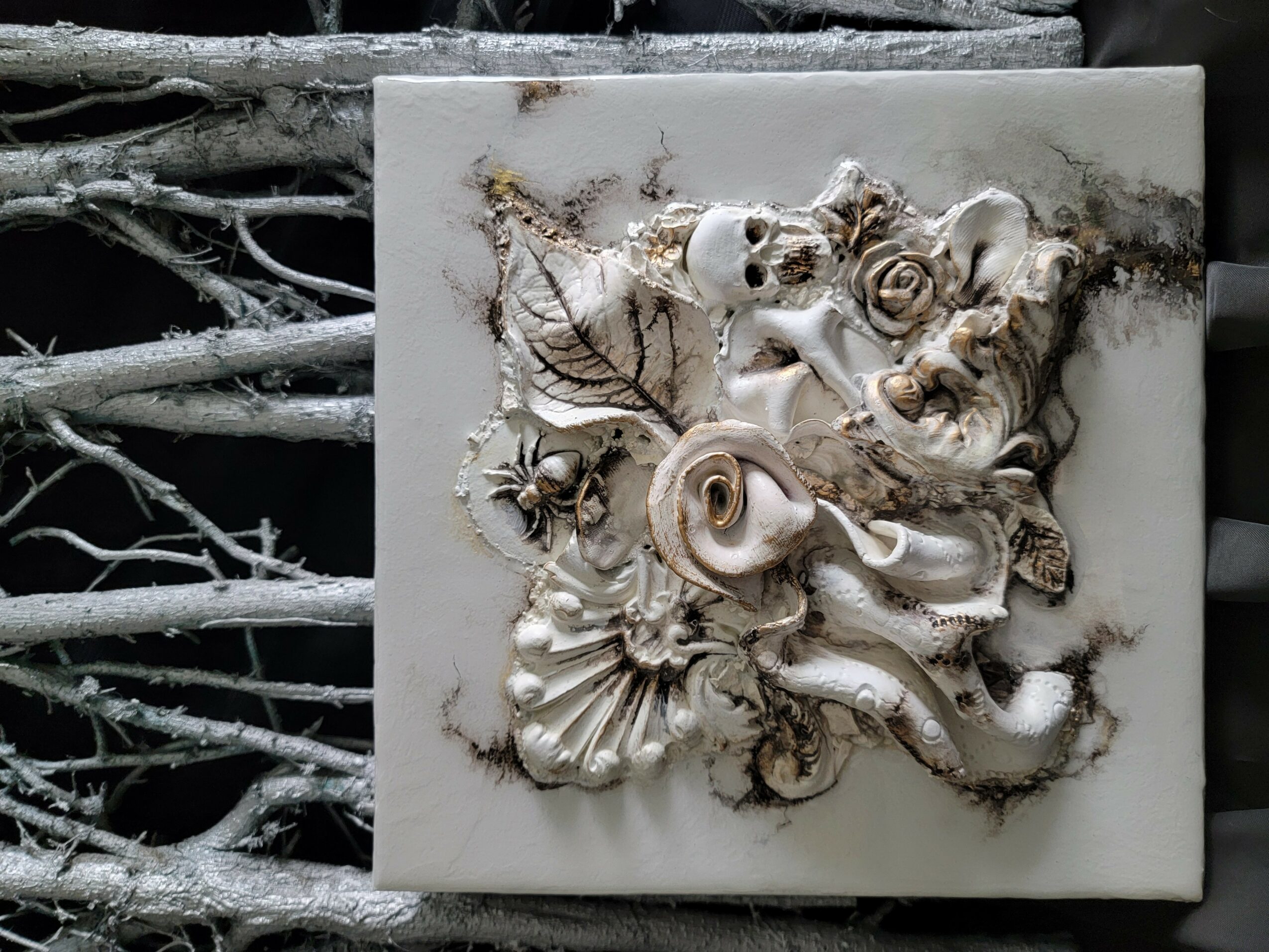 Decorative relief with flowers, leaves, and a face, mounted on a wall with branches in the background.