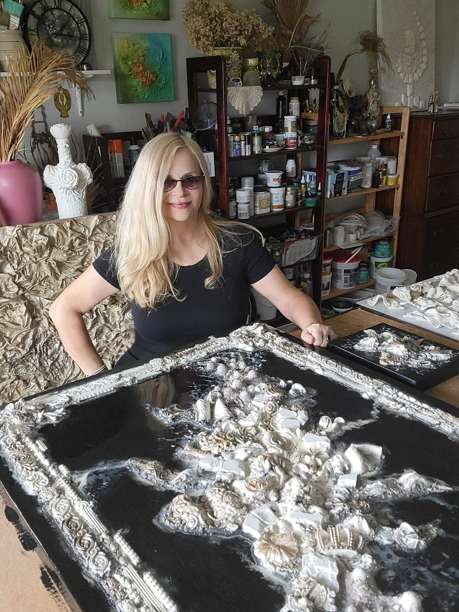 Woman with long blonde hair and sunglasses standing behind a tray of jewelry or metal objects, in a room with shelves and artwork.
