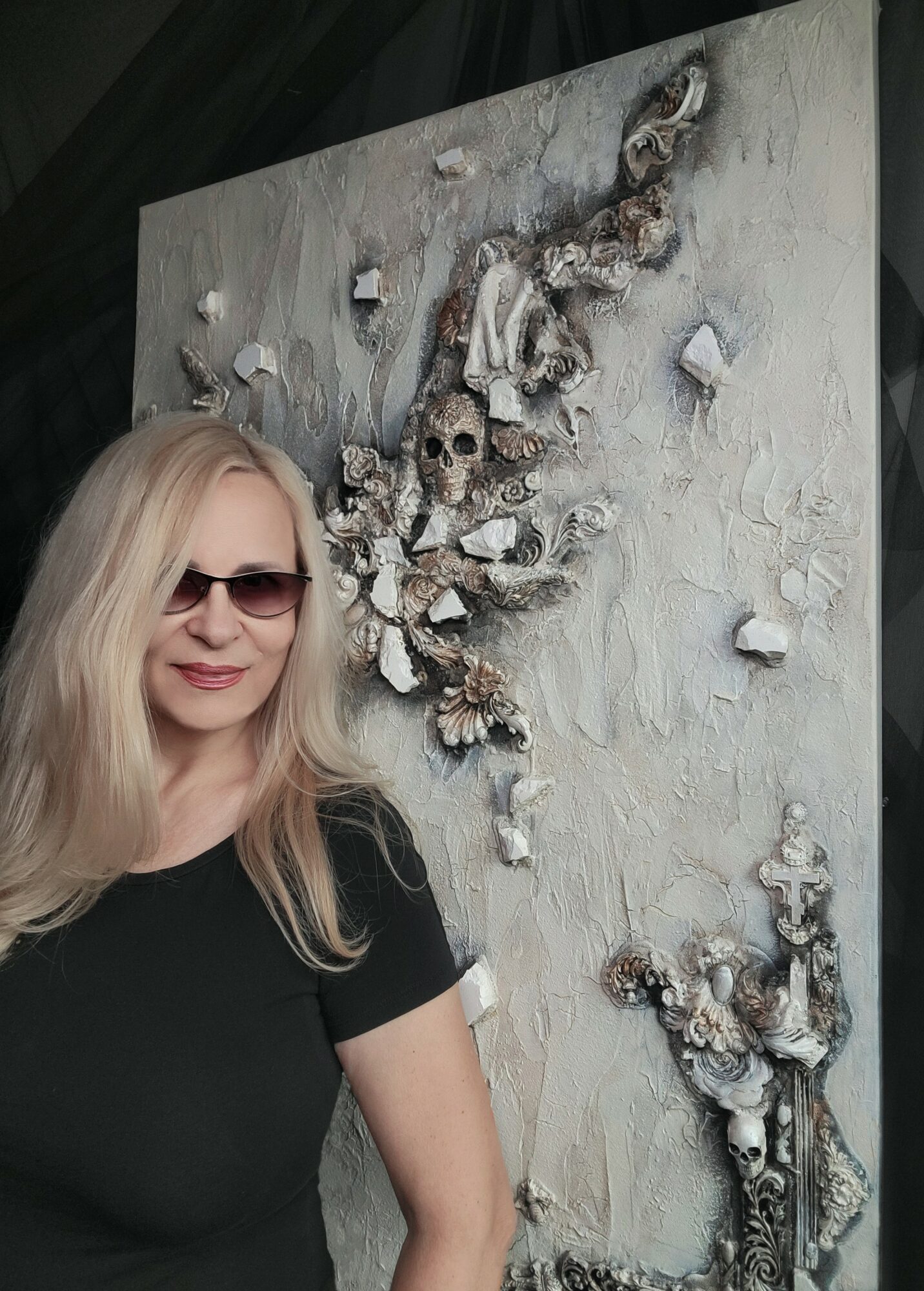 Woman with long blonde hair and sunglasses standing next to a textured artwork with skulls and shells.