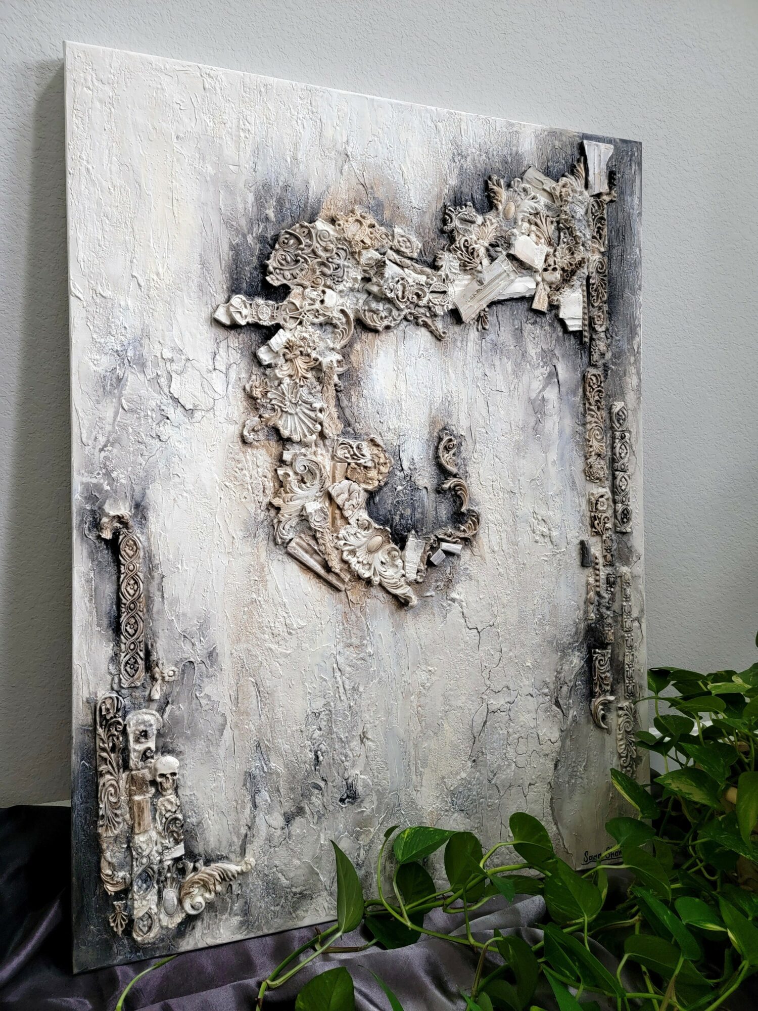 Mixed media artwork with textured, layered elements forming a heart shape on a gray background, with green plants in foreground.