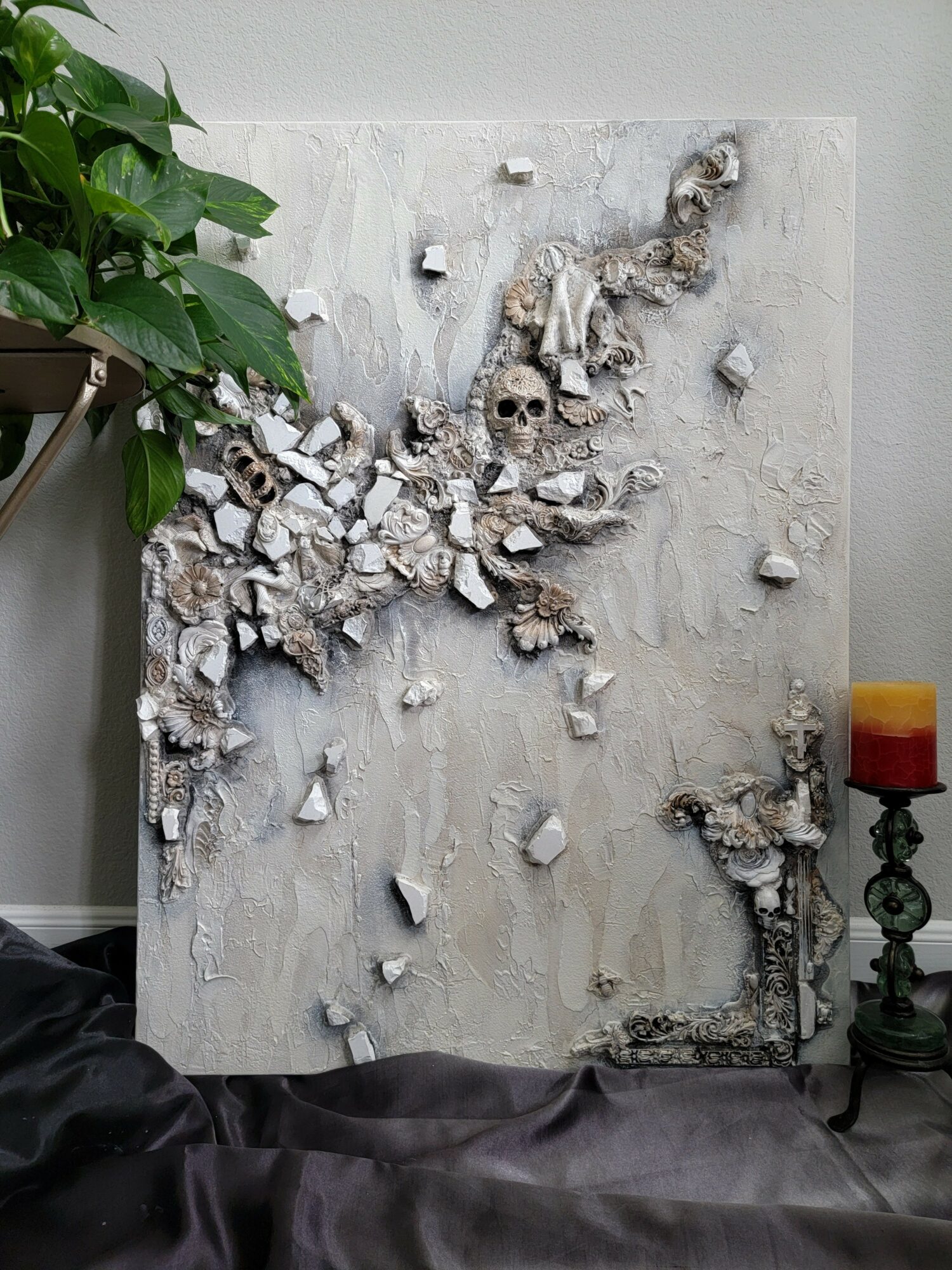 Abstract textured artwork with white, beige, and brown elements on a light background, next to a green plant and a candle holder.