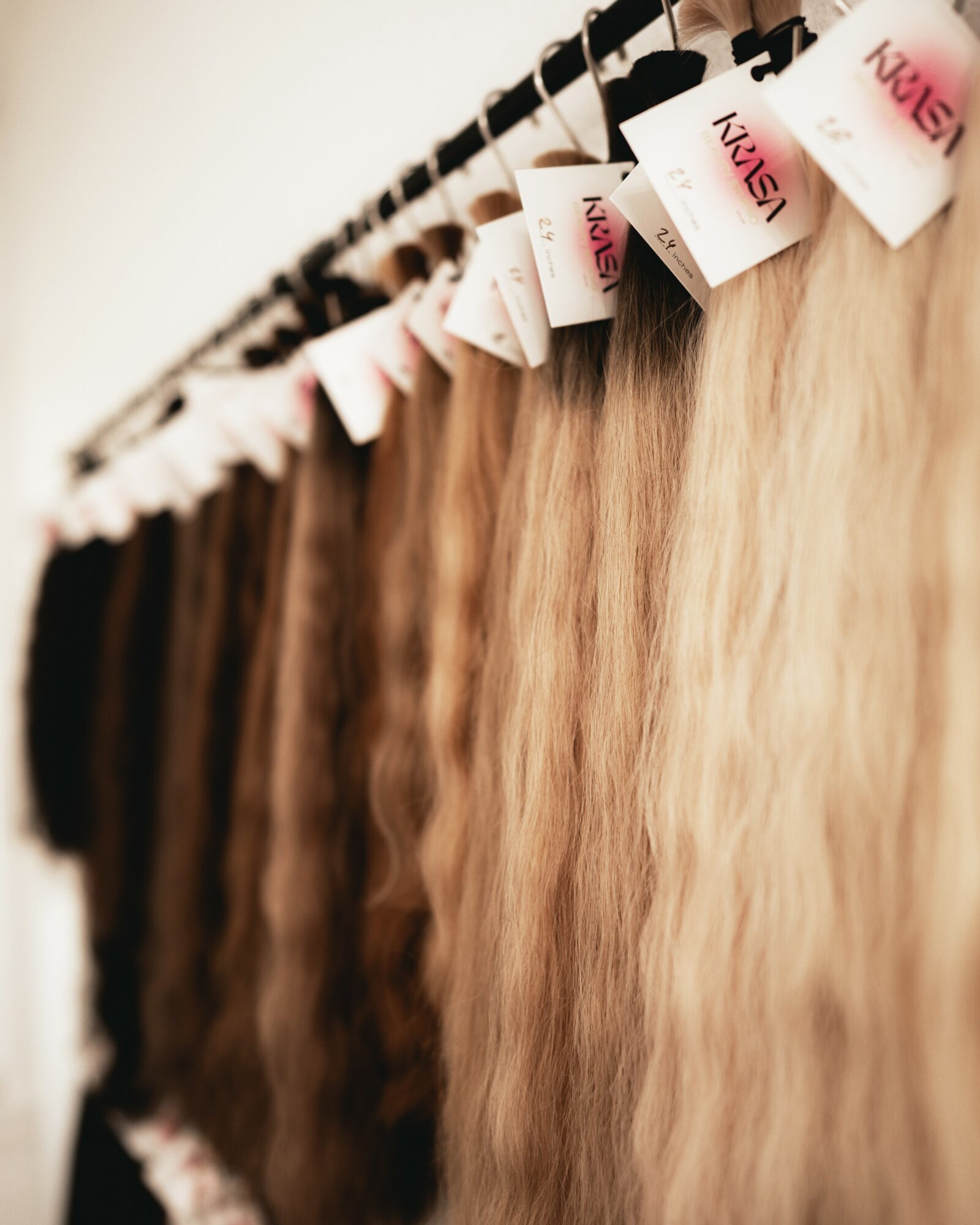 Multiple hair extensions hanging on a rack, varying shades from dark to light blonde, with tags attached.
