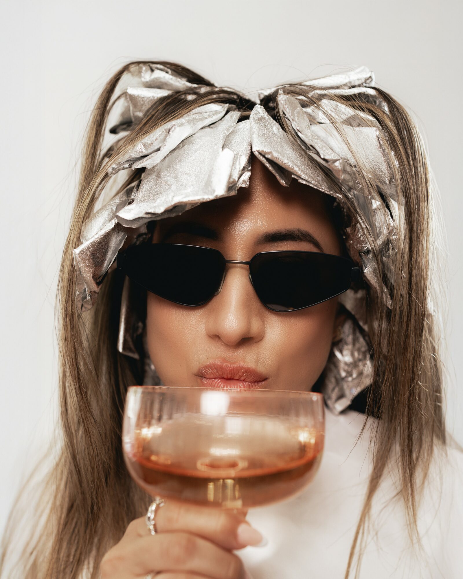 Person wearing sunglasses and a large bow headband, holding a glass with a drink, neutral background.