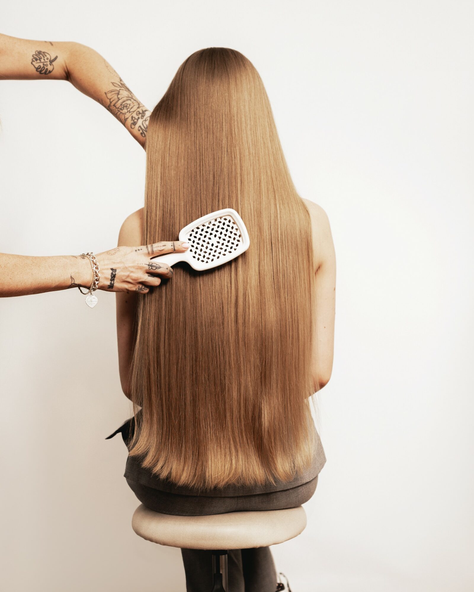 Person with long, straight, light brown hair sitting on a stool, brushing hair with a white hairbrush, against a plain white background.