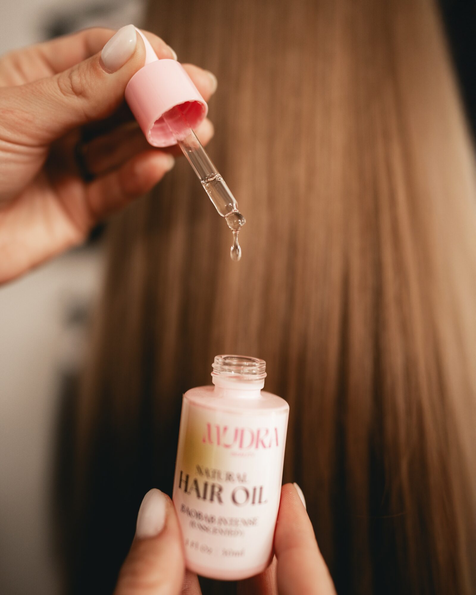 Person holding a dropper above a bottle of hair oil, with hair in the background.