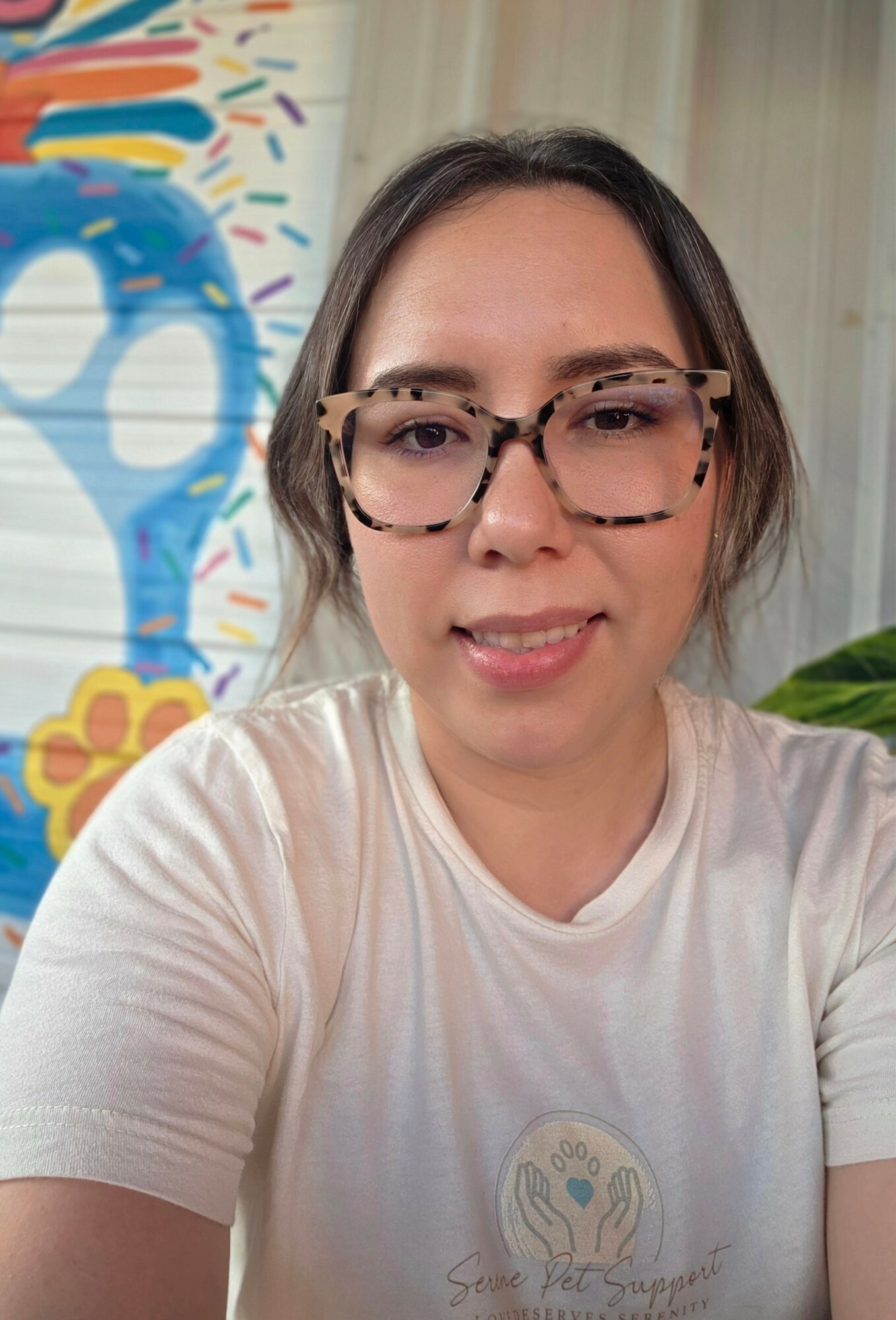 Woman with glasses and dark hair in a white shirt, smiling indoors with colorful wall art behind her.