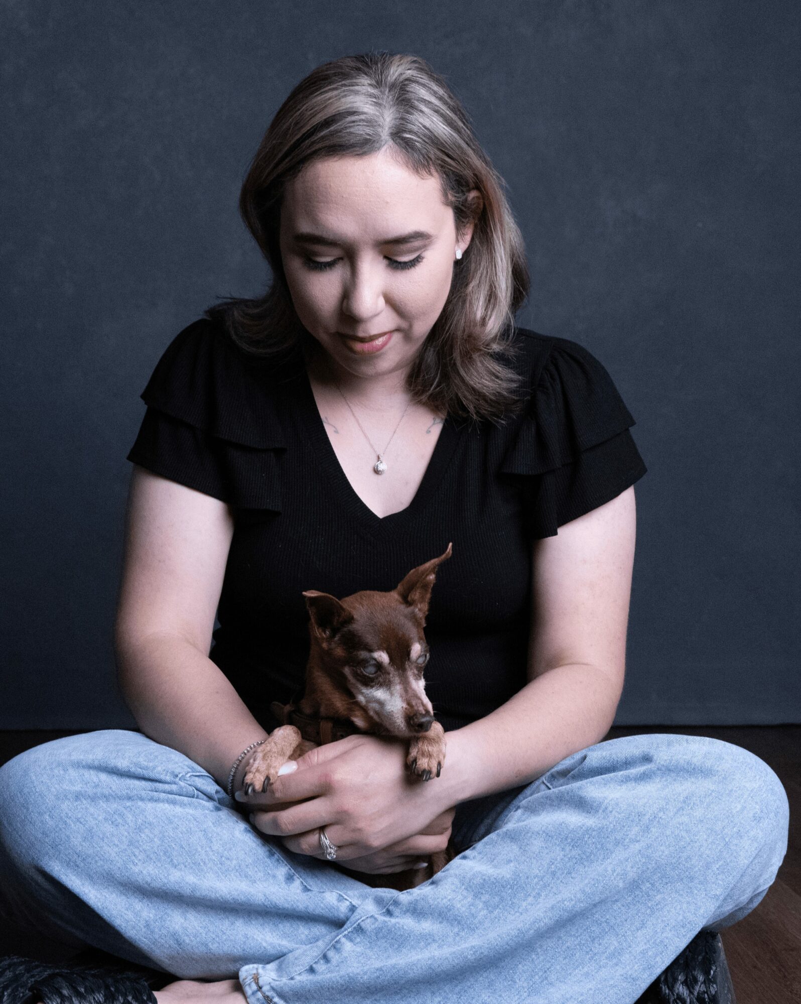 Woman sitting cross-legged holding a small brown dog against a dark background.