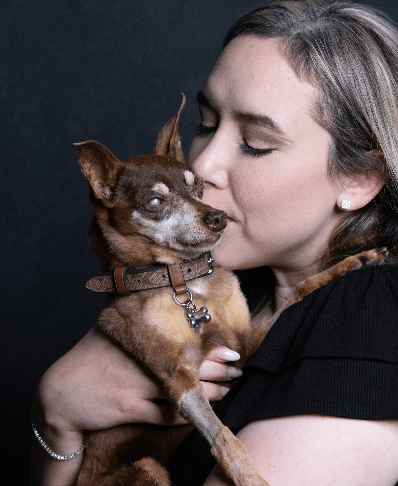 Woman holding and kissing a small dog with a brown coat and white markings, against a dark background.