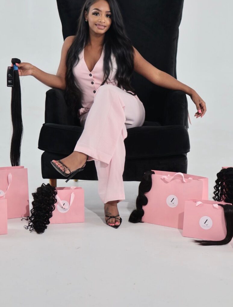 Woman sitting on a black throne-like chair, surrounded by pink gift bags and black hair extensions, smiling.