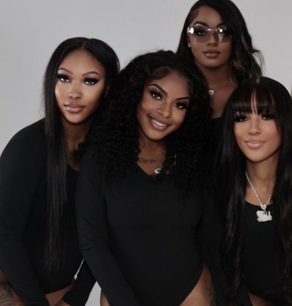 Four women with dark hair and makeup pose together against a plain background, smiling and dressed in black. Word count: 20
