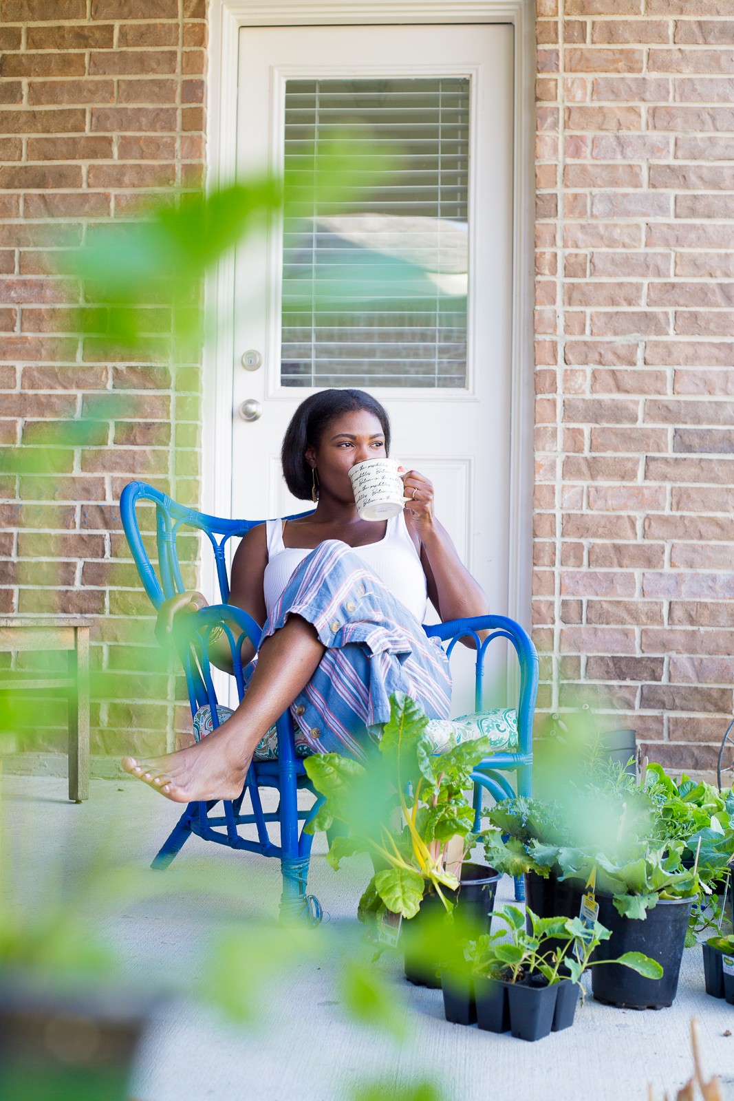 Girl sitting on a blue chair outside, drinking from a cup, with a door and brick wall behind her.