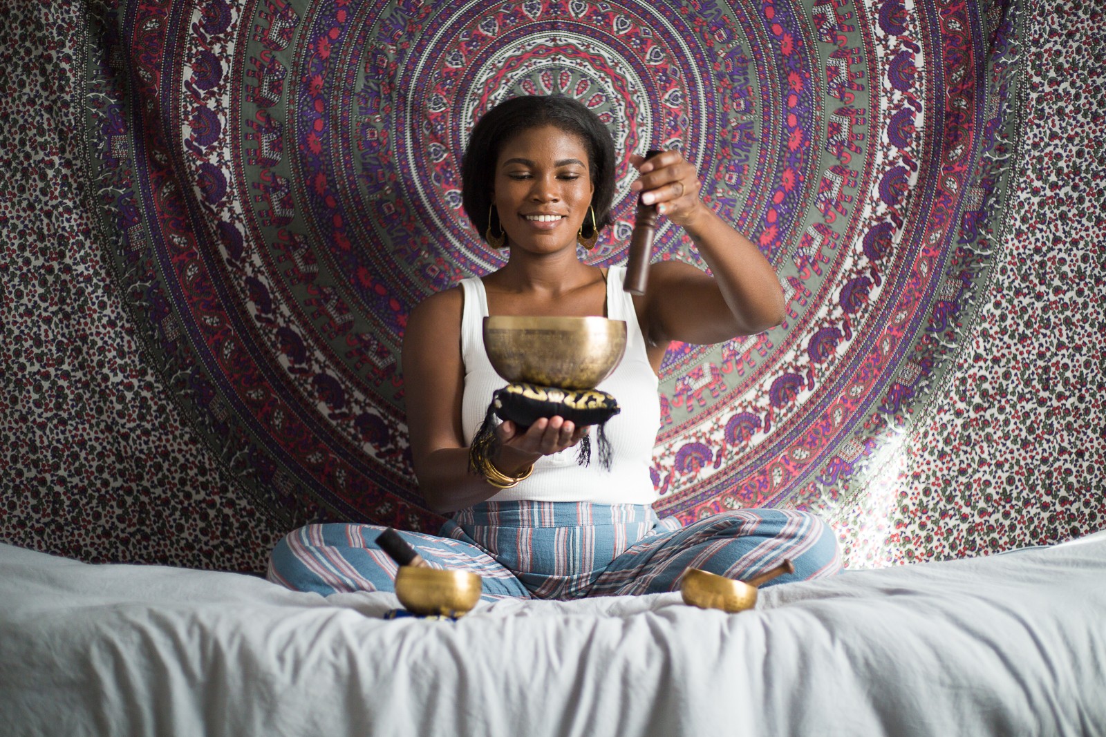 Woman sitting cross-legged on bed, holding a singing bowl, smiling, with a colorful tapestry background.