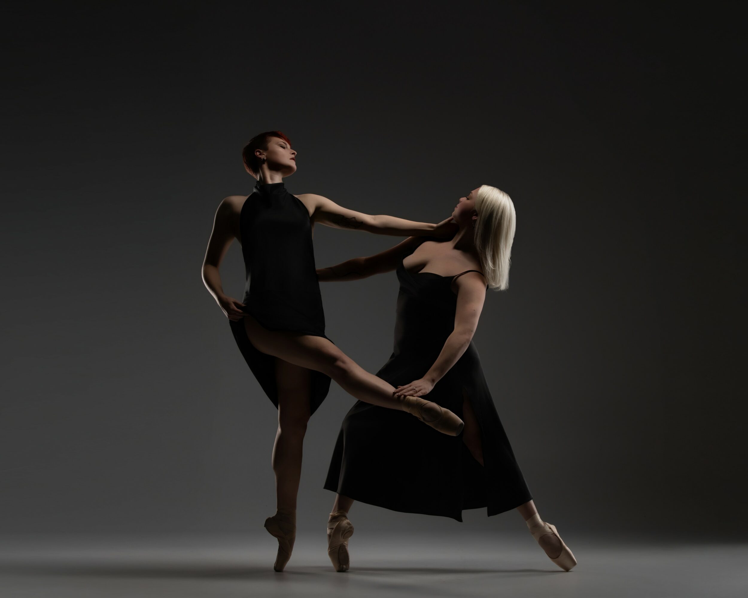 Two ballet dancers, one with dark clothing and one with light hair, perform a pose against a dark background.
