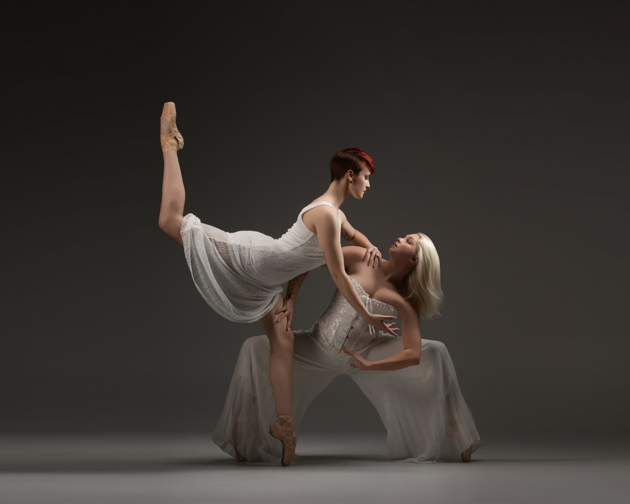 Two ballet dancers, one standing on pointe with one leg raised, the other kneeling, holding and supporting each other, against a dark background.