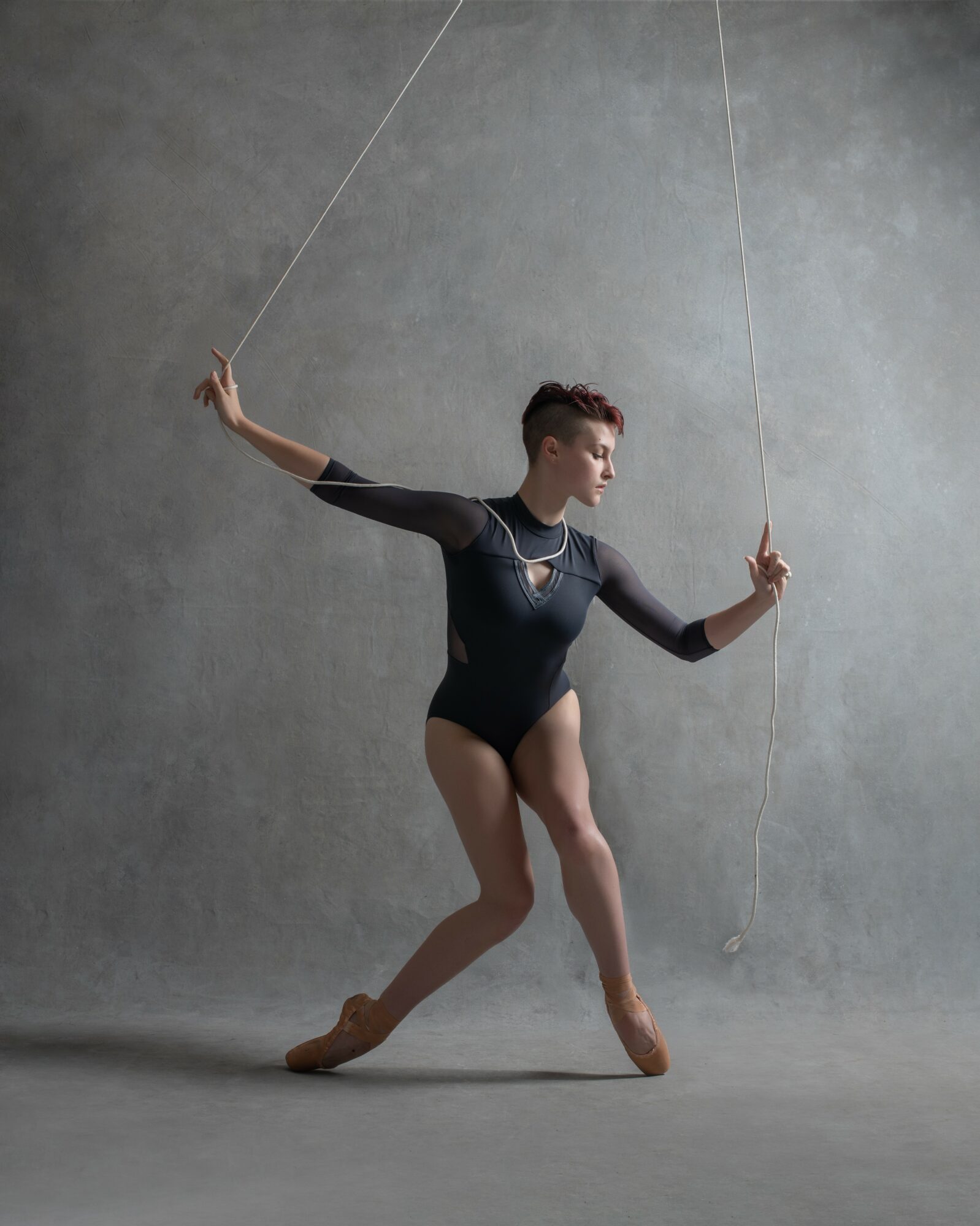 Ballet dancer in black leotard poses with arms extended, holding strings against a gray background.