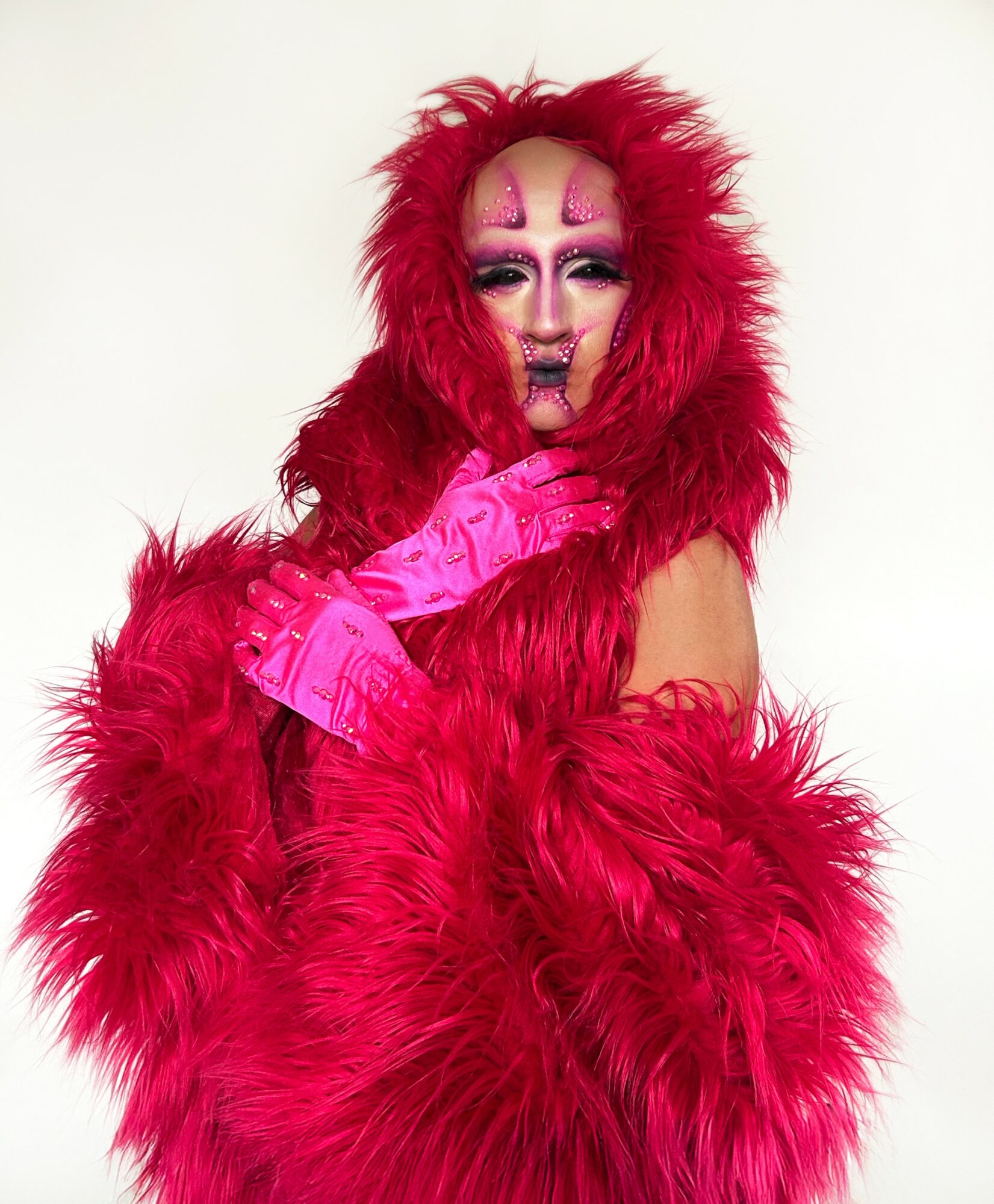 Person in elaborate pink costume with feathered coat, makeup, and gloves, posing with hand near face against a white background.
