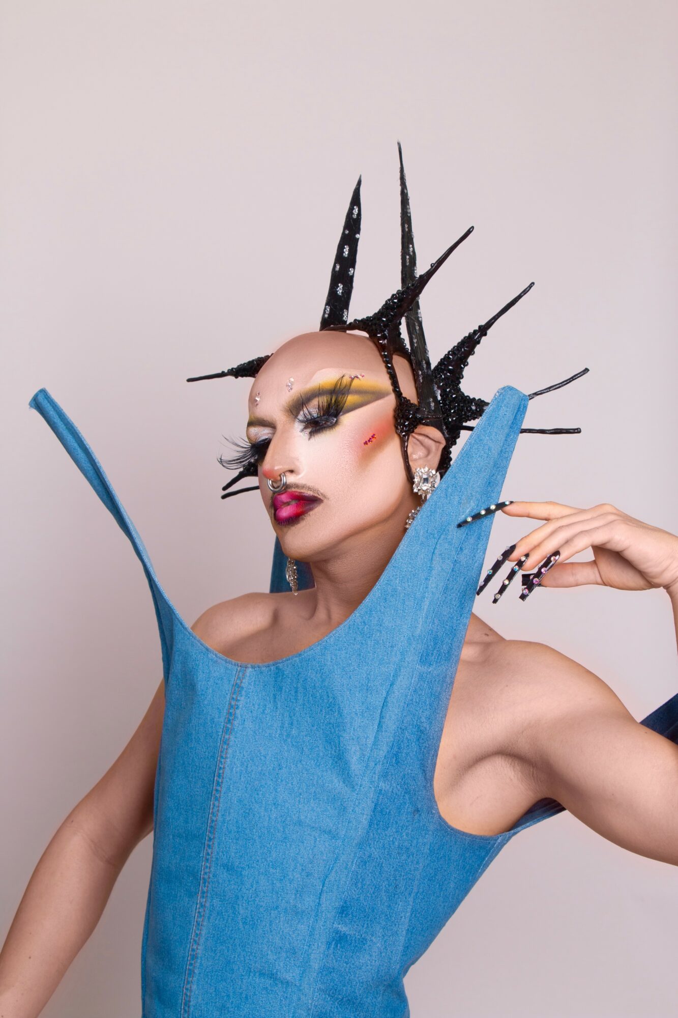 Person with elaborate black spiked headpiece and colorful makeup, wearing a blue garment with a high collar, posing with hand near face.