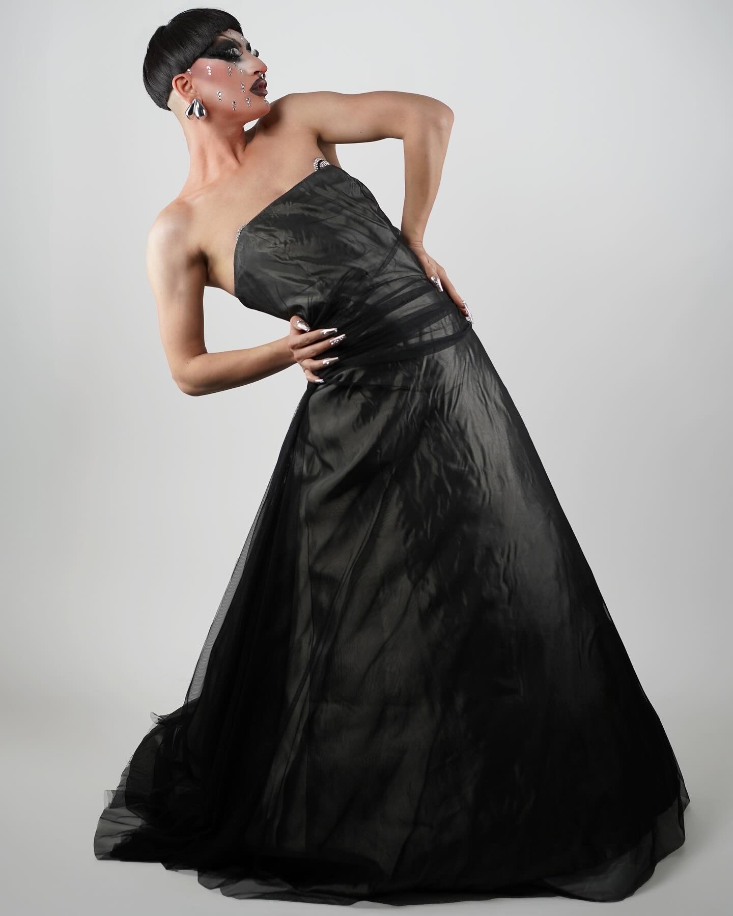 Woman in a strapless black gown with short hair and glasses, posing with one hand on her hip, against a plain background.