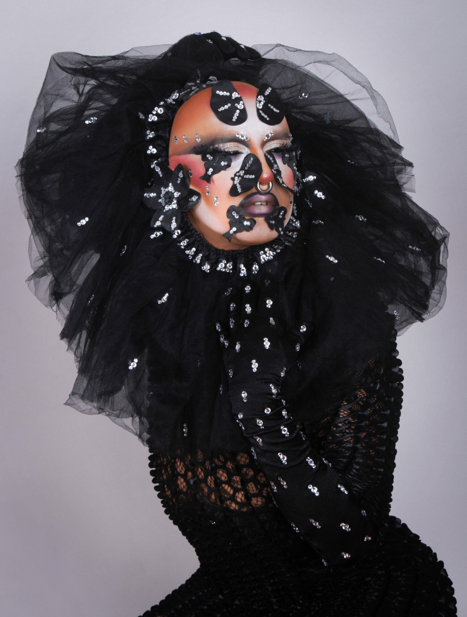 Person with elaborate makeup and black tulle headpiece, wearing black lace clothing, facing sideways with hand near face.