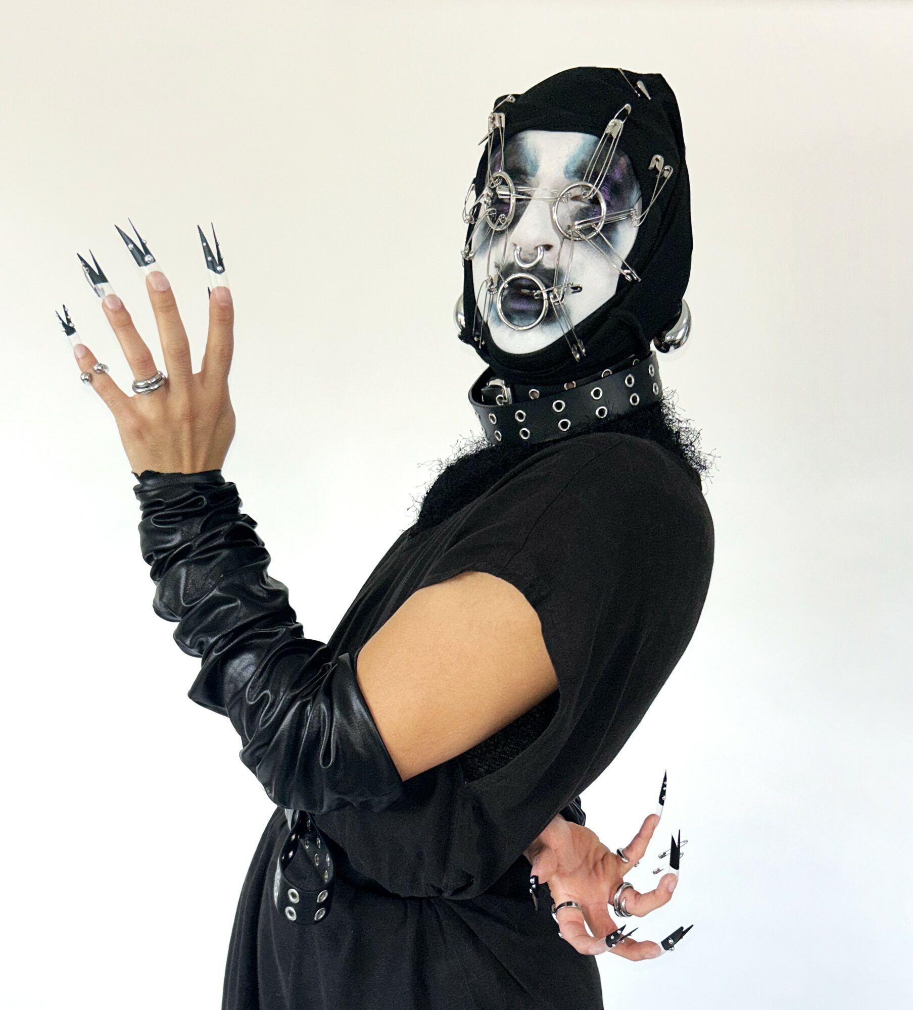 Person with a mask, glasses, and black clothing, holding up a hand with long, pointed nails, against a plain background.
