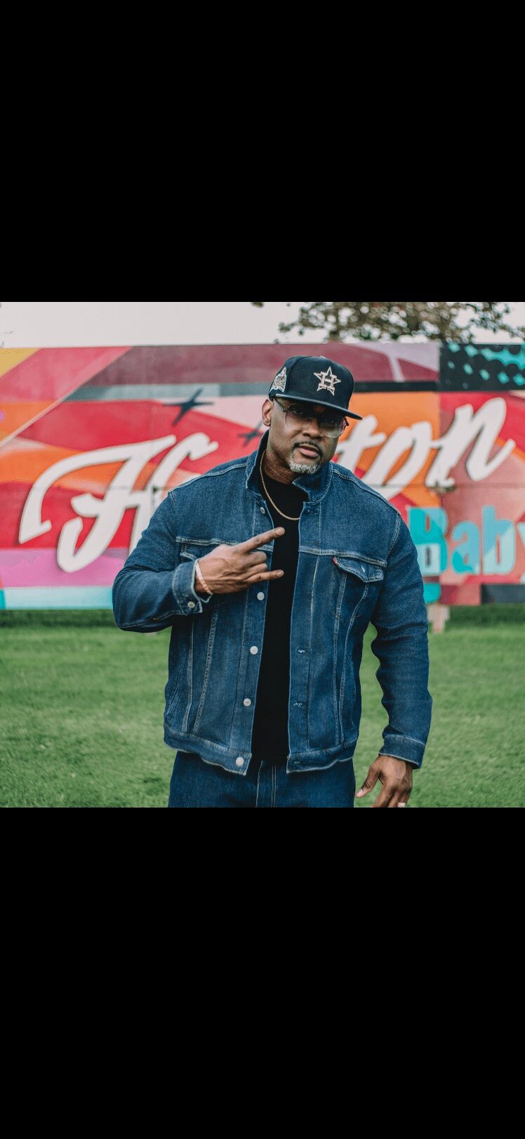 Man wearing a denim jacket and cap, standing outdoors in front of a colorful Ford sign.