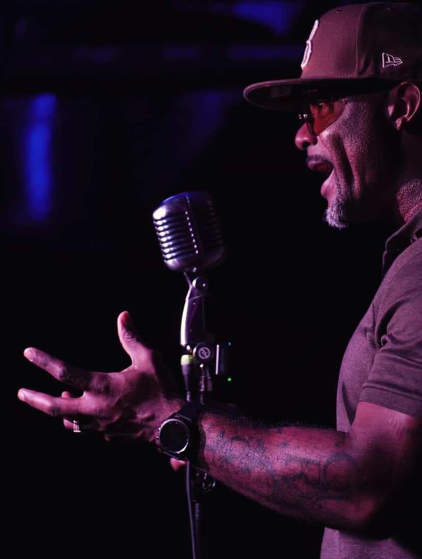 Man wearing a cap and glasses speaking into a microphone, gesturing with his hand, in a dark setting.