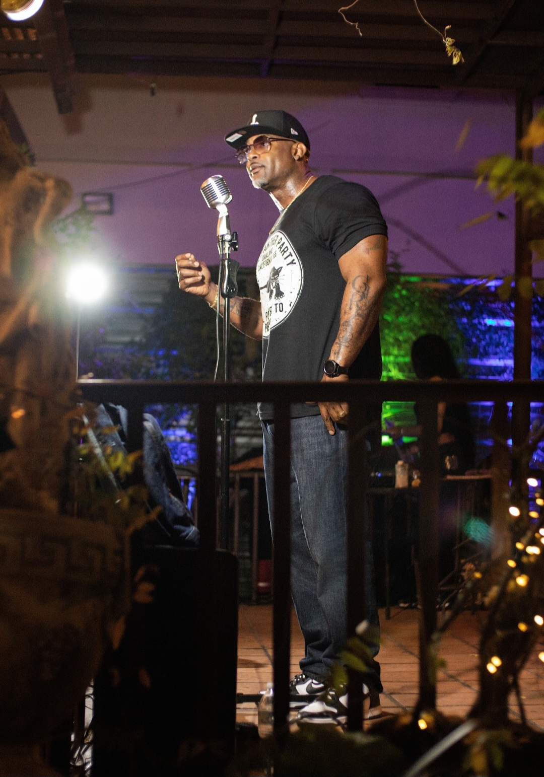 Man in black shirt and cap speaking into microphone on stage with colorful lights, holding a drink, in a dimly lit venue.