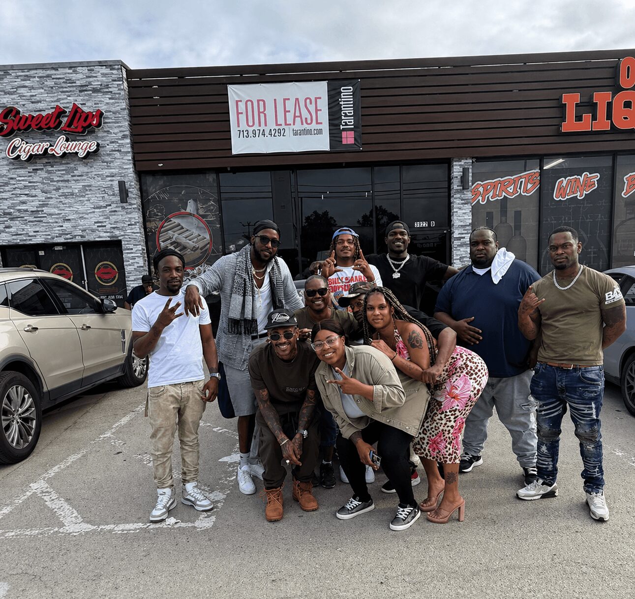 Group of nine people standing outside a building with a sign for lease, some making peace signs, in front of parked cars.