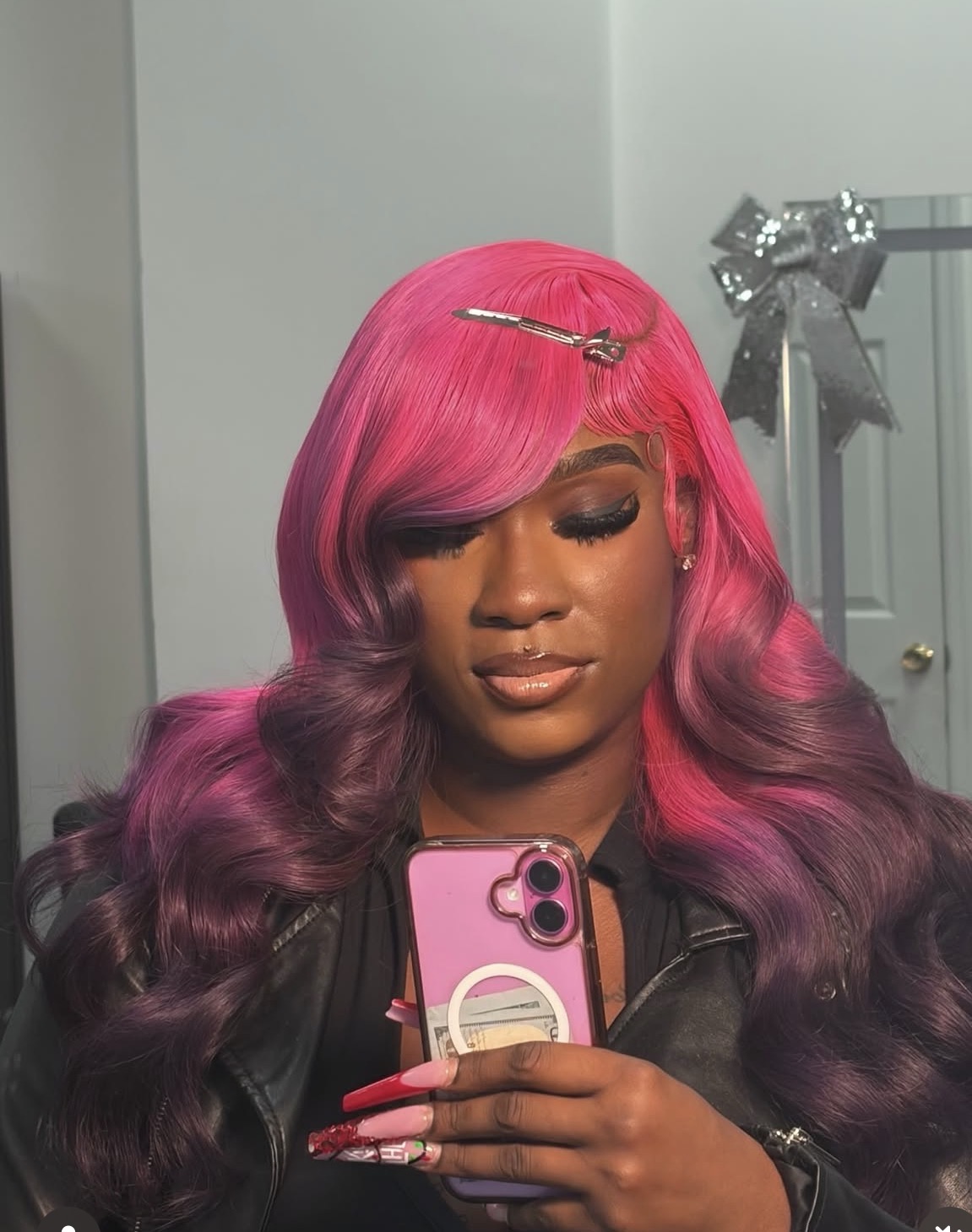 Woman with pink and purple wavy hair taking a selfie with a pink phone, wearing makeup and a black jacket.