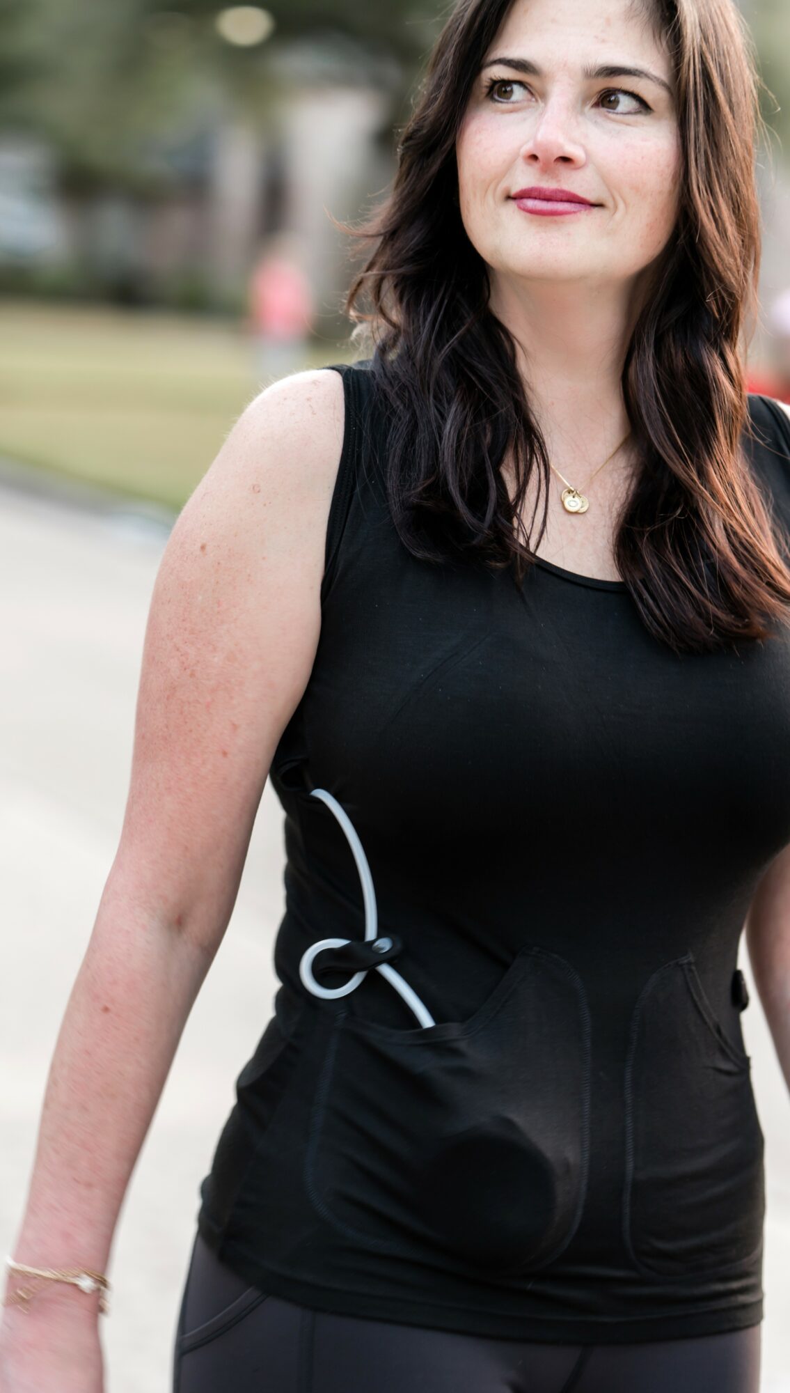 Woman with long dark hair wearing a black sleeveless top outdoors, looking to the side, smiling.
