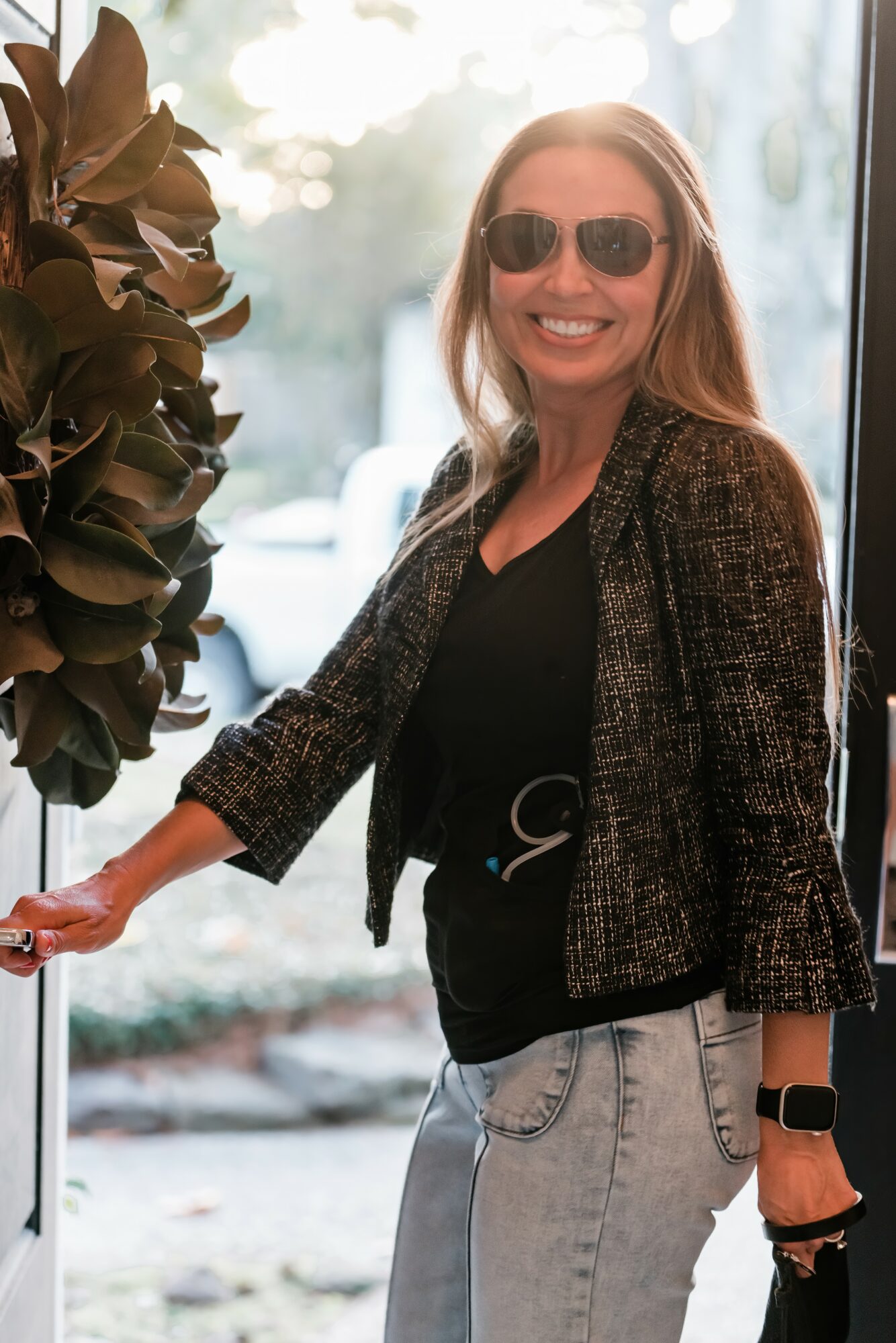 Woman smiling wearing sunglasses, black top, patterned blazer, and light jeans, standing near a window with plants outside.