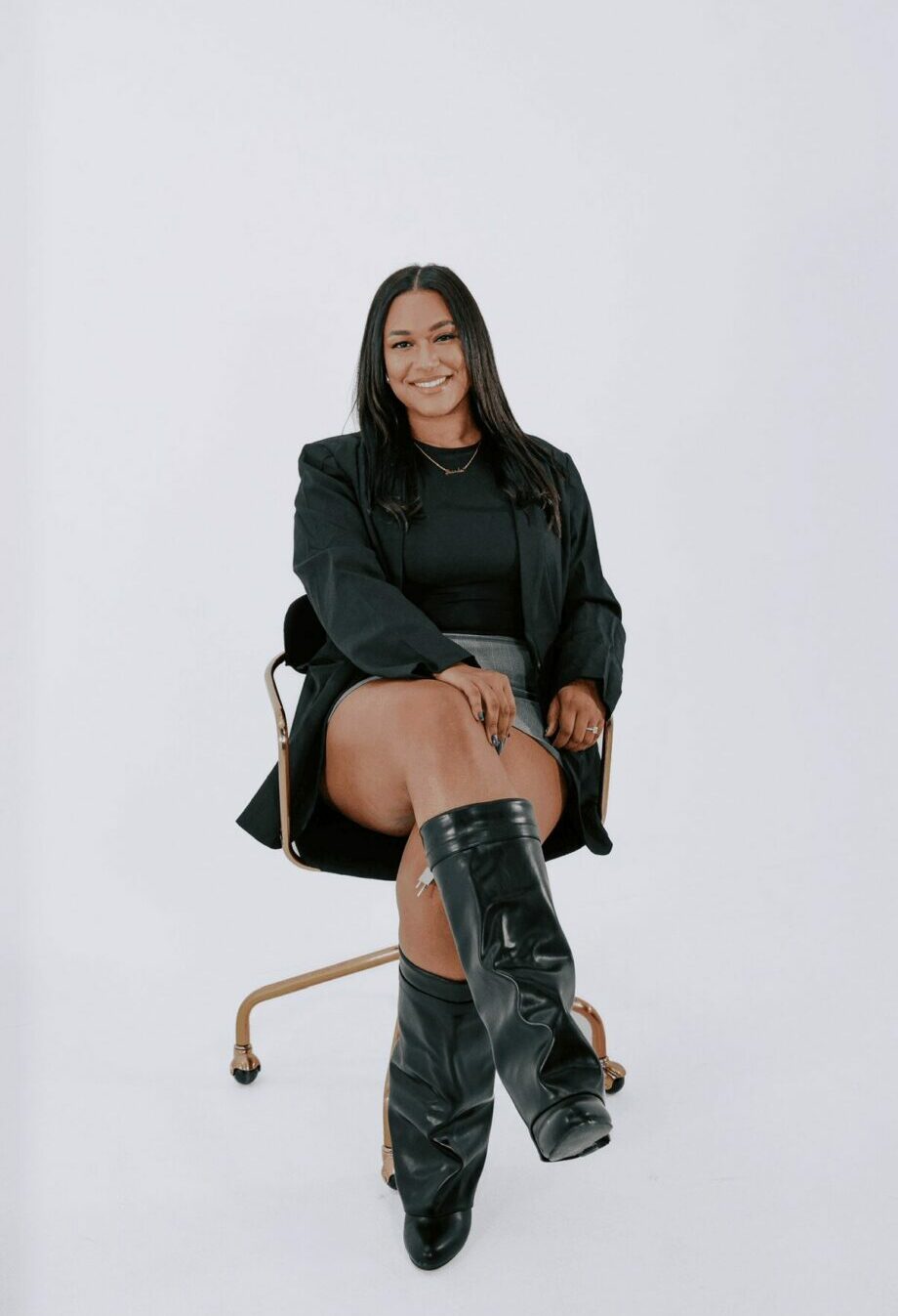 Woman sitting on a chair with a white background, smiling, wearing a black outfit and thigh-high boots.