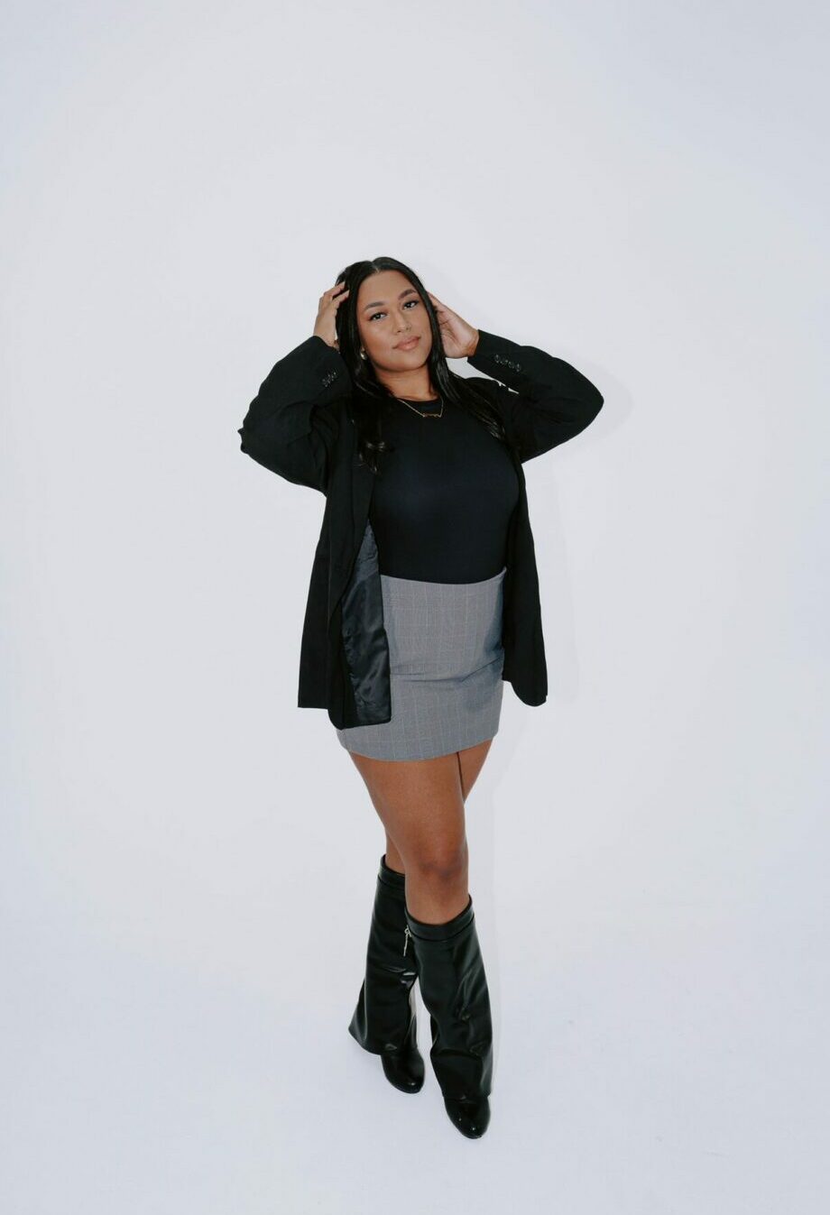 Woman standing with hands near her head, wearing a black jacket, black top, gray skirt, and black boots, against a white background.