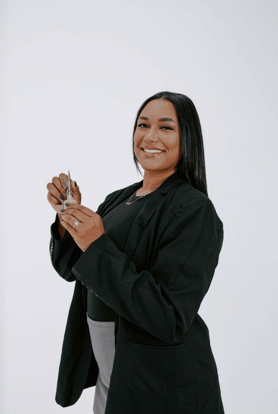 Smiling woman with long dark hair wearing a black blazer and gray pants, standing against a white background.