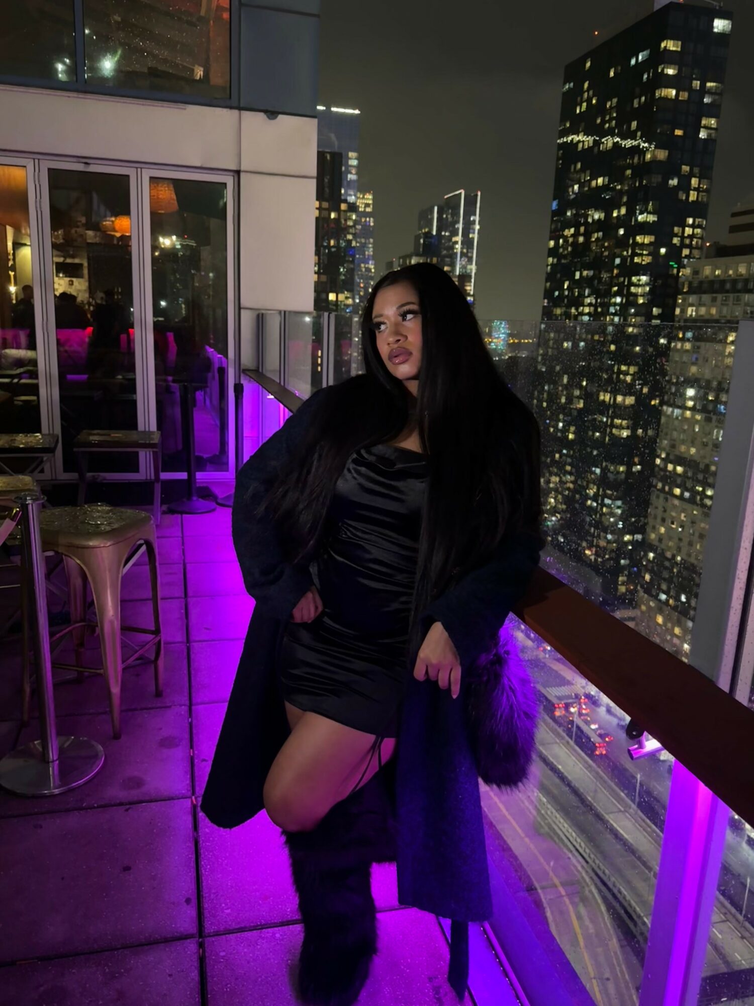 Woman standing on a balcony at night with city skyline and illuminated buildings behind her.
