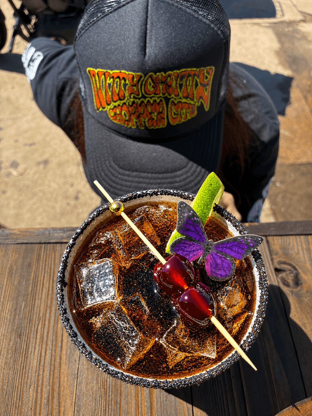 Top view of a drink with ice, garnished with a lime wedge, purple butterfly, and cherry, on a wooden surface.