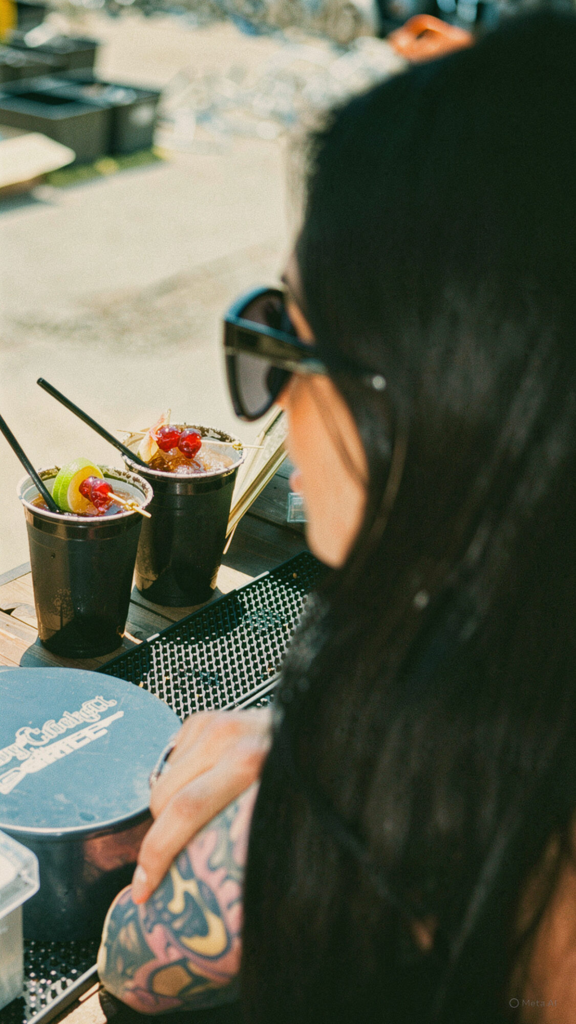 Person with dark hair and tattooed arm sitting at a table with three drinks topped with fruit, outdoors.