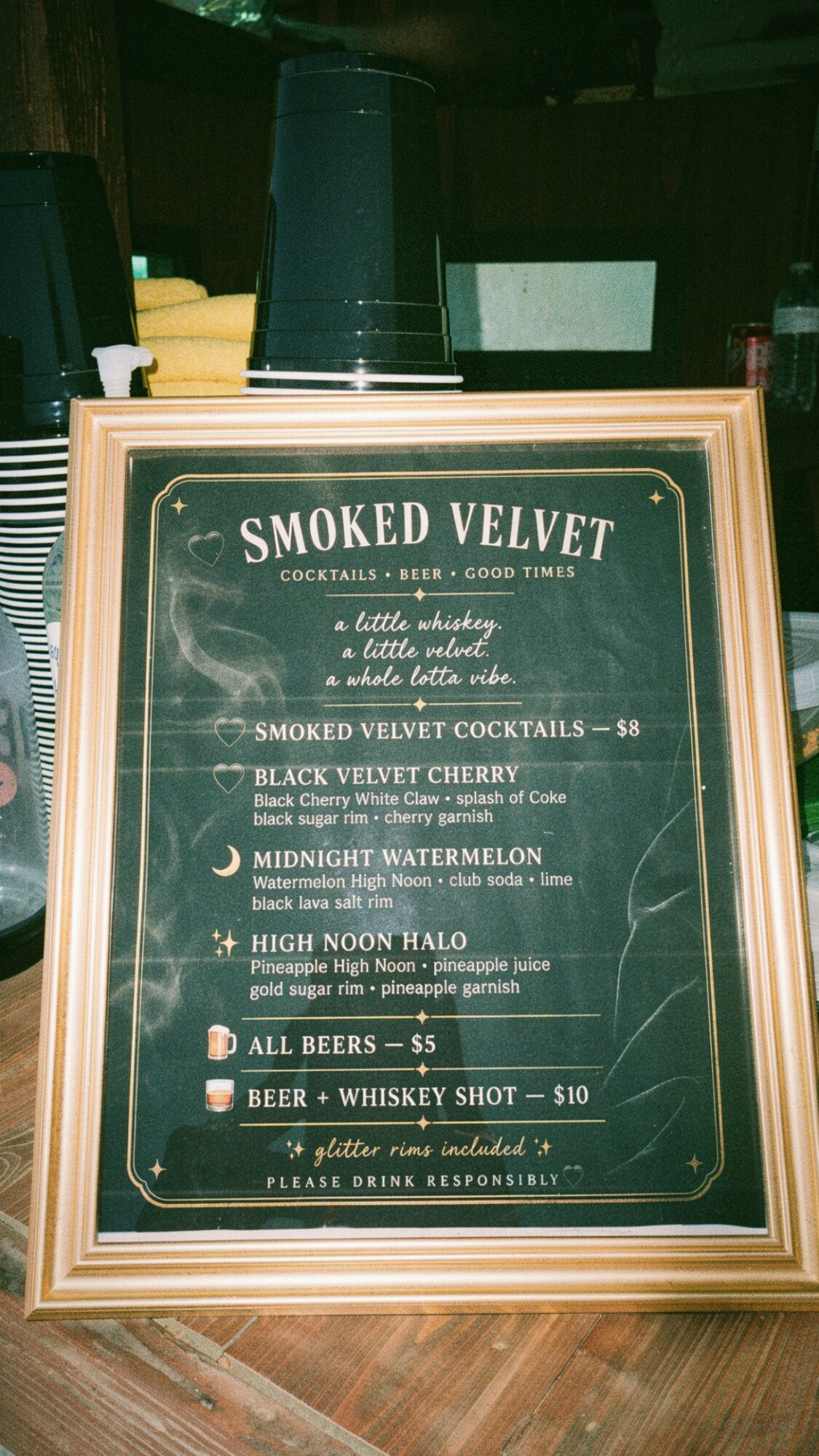 Menu titled 'Smoked Velvet' with drink options and prices, framed on a wooden surface, with cups and bottles in background.