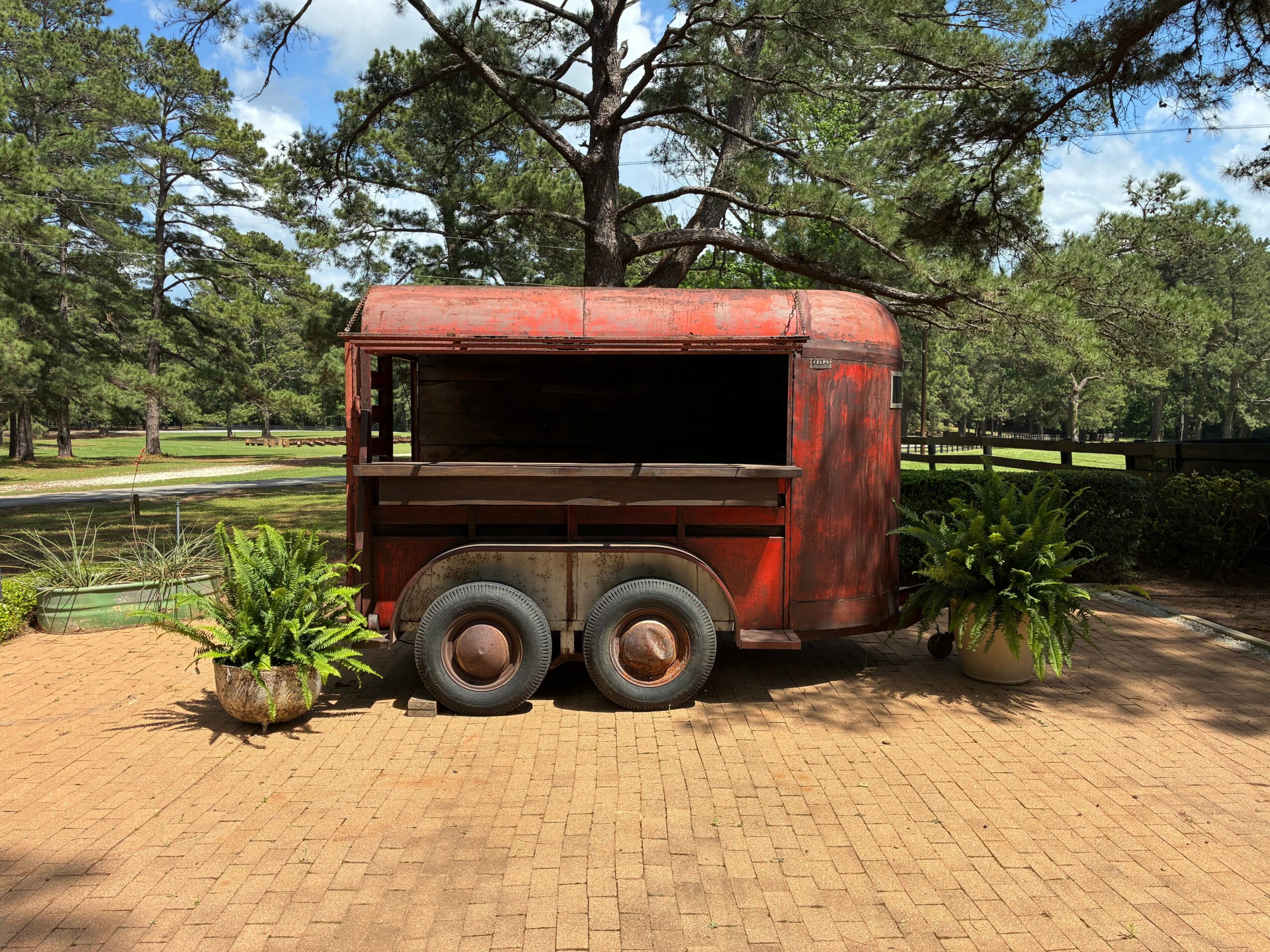 Rusty food truck with two potted plants on brick ground, trees and a fence in background.