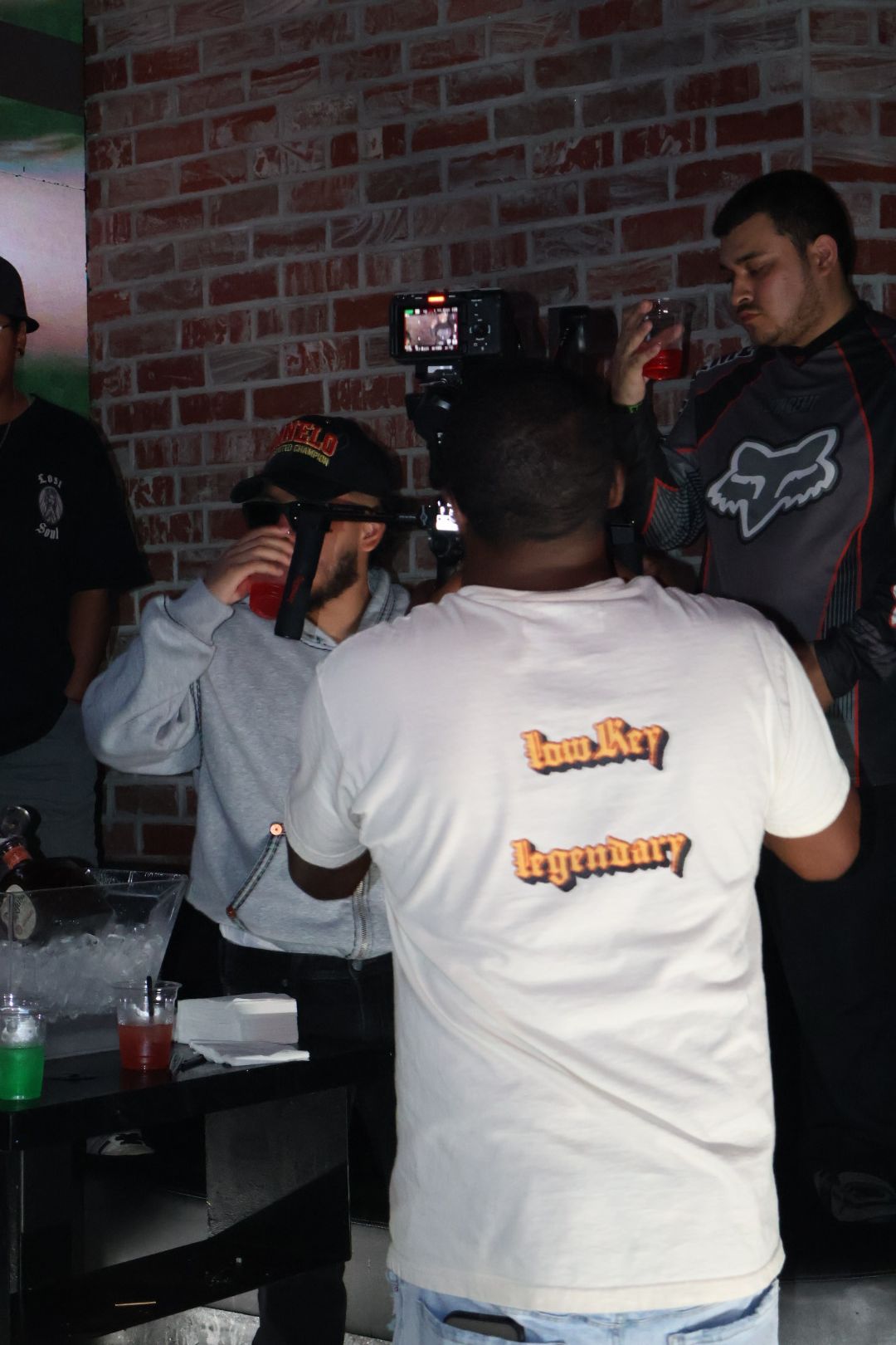 Person with short hair wearing a white T-shirt with text, sitting at a table with a brick wall background.