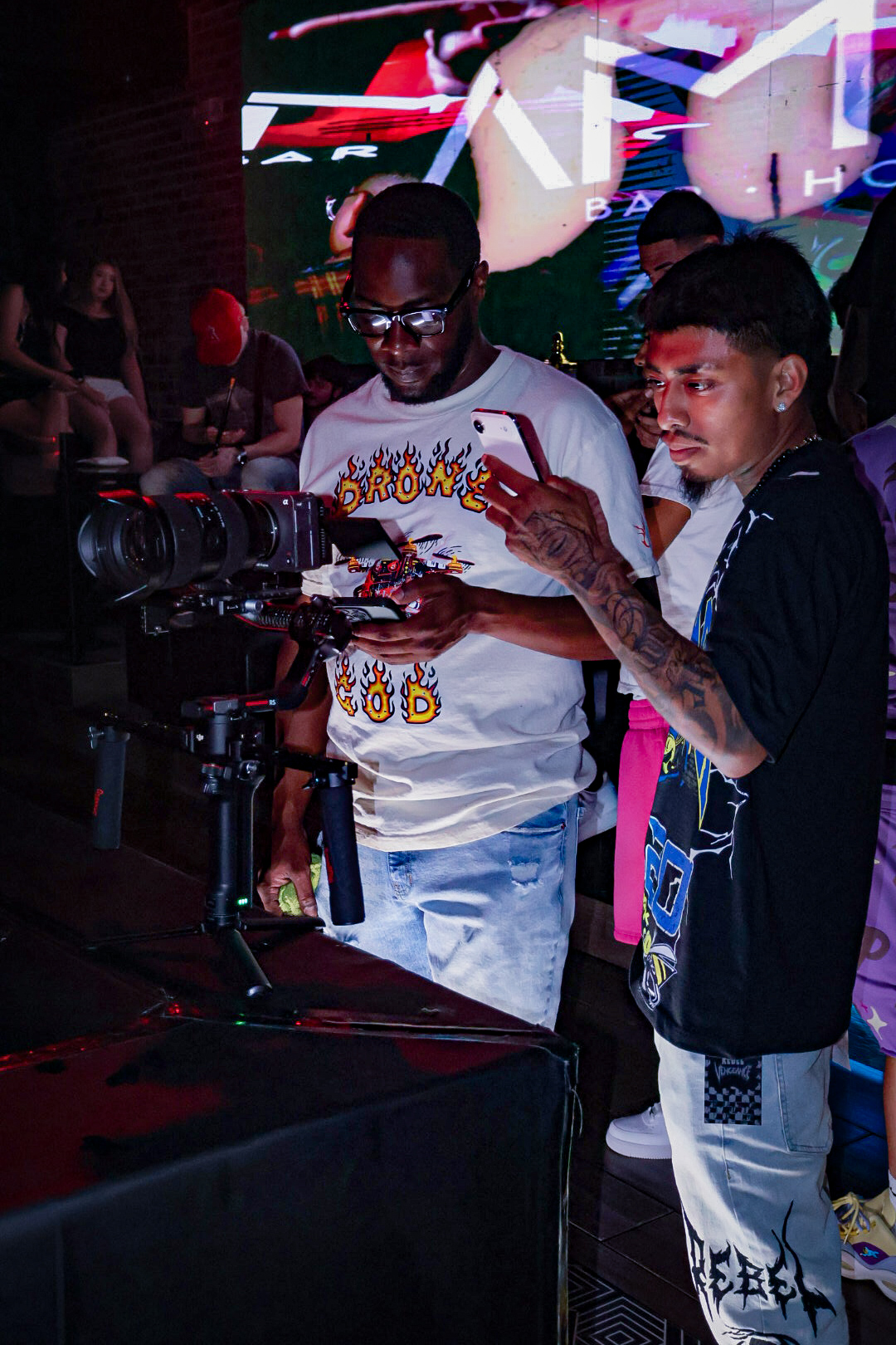 Two men look at a camera setup in a dark environment with colorful lights and a large screen in the background.