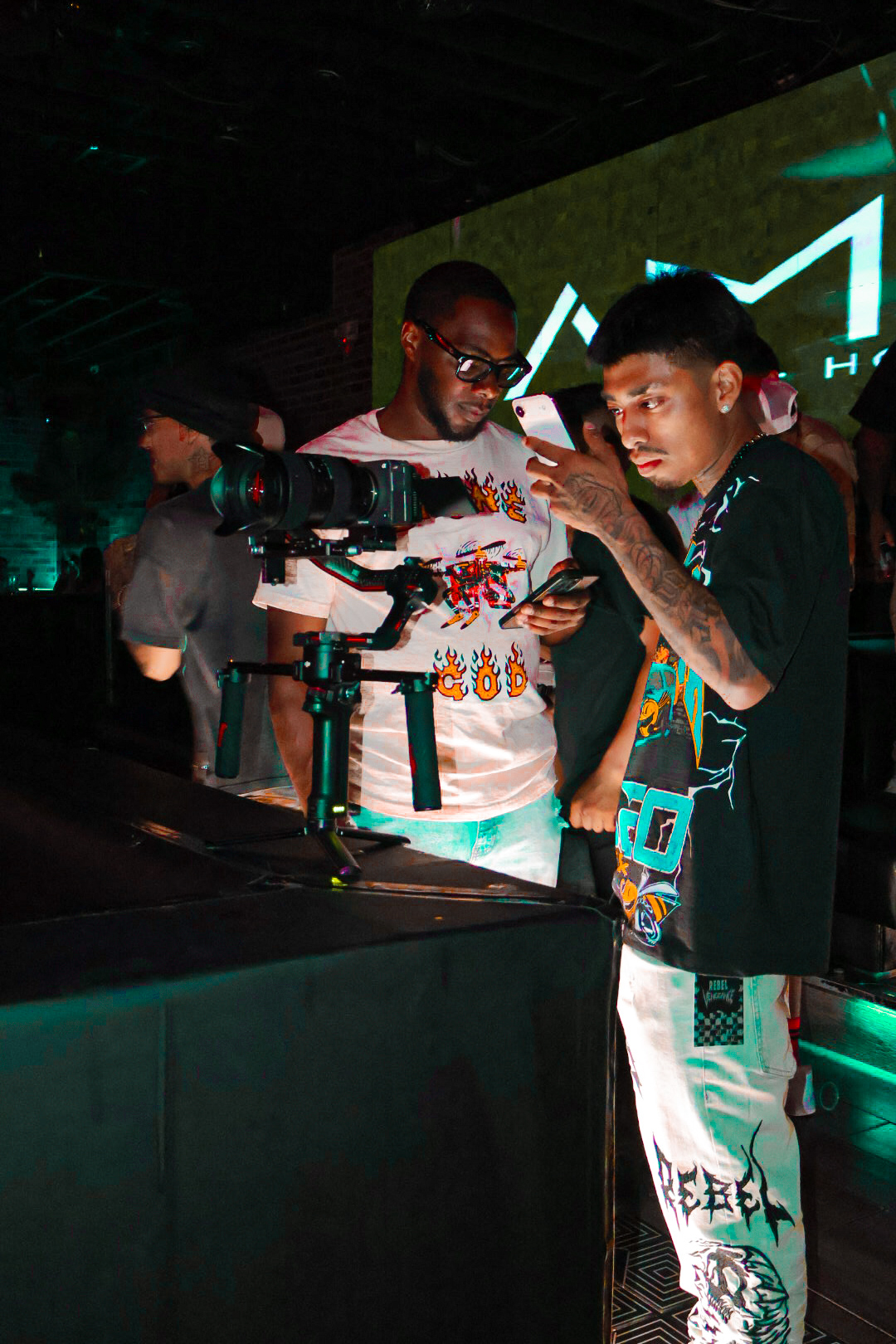 Two young men are examining a camera setup at an event with a large screen in the background, dim lighting, and a dark environment.