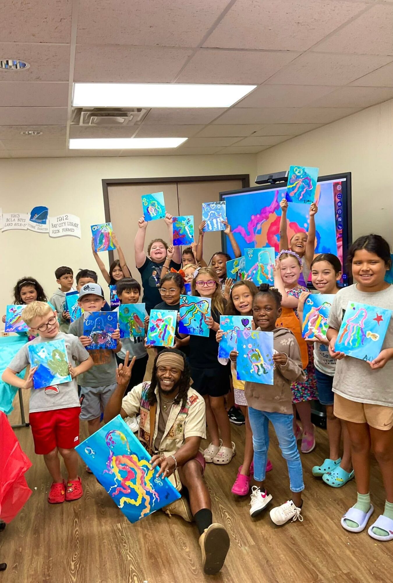 Group of children and an adult in a classroom holding colorful paintings, some raising their artwork, with a large screen in the background.