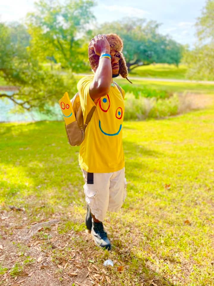 Child walking outdoors on grass with trees and a pond in the background, wearing a yellow shirt with smiley face, carrying a backpack.