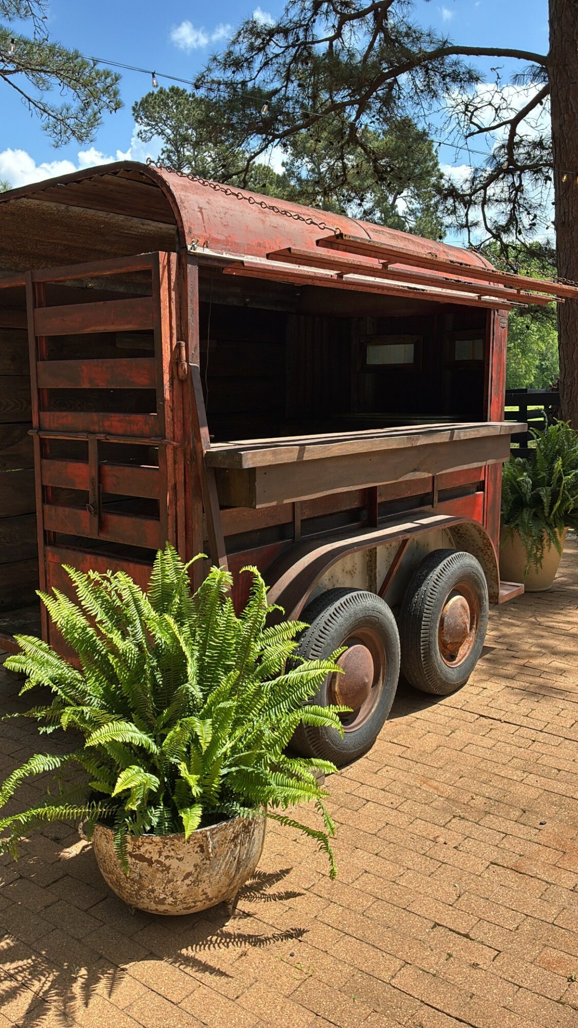 Wooden cart with wheels and a red roof, surrounded by green plants and trees, on a brick surface.
