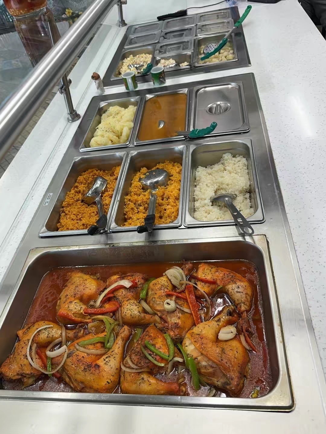 Tray of cooked chicken with vegetables and sauce, various toppings, and condiments in metal containers, on a white counter.