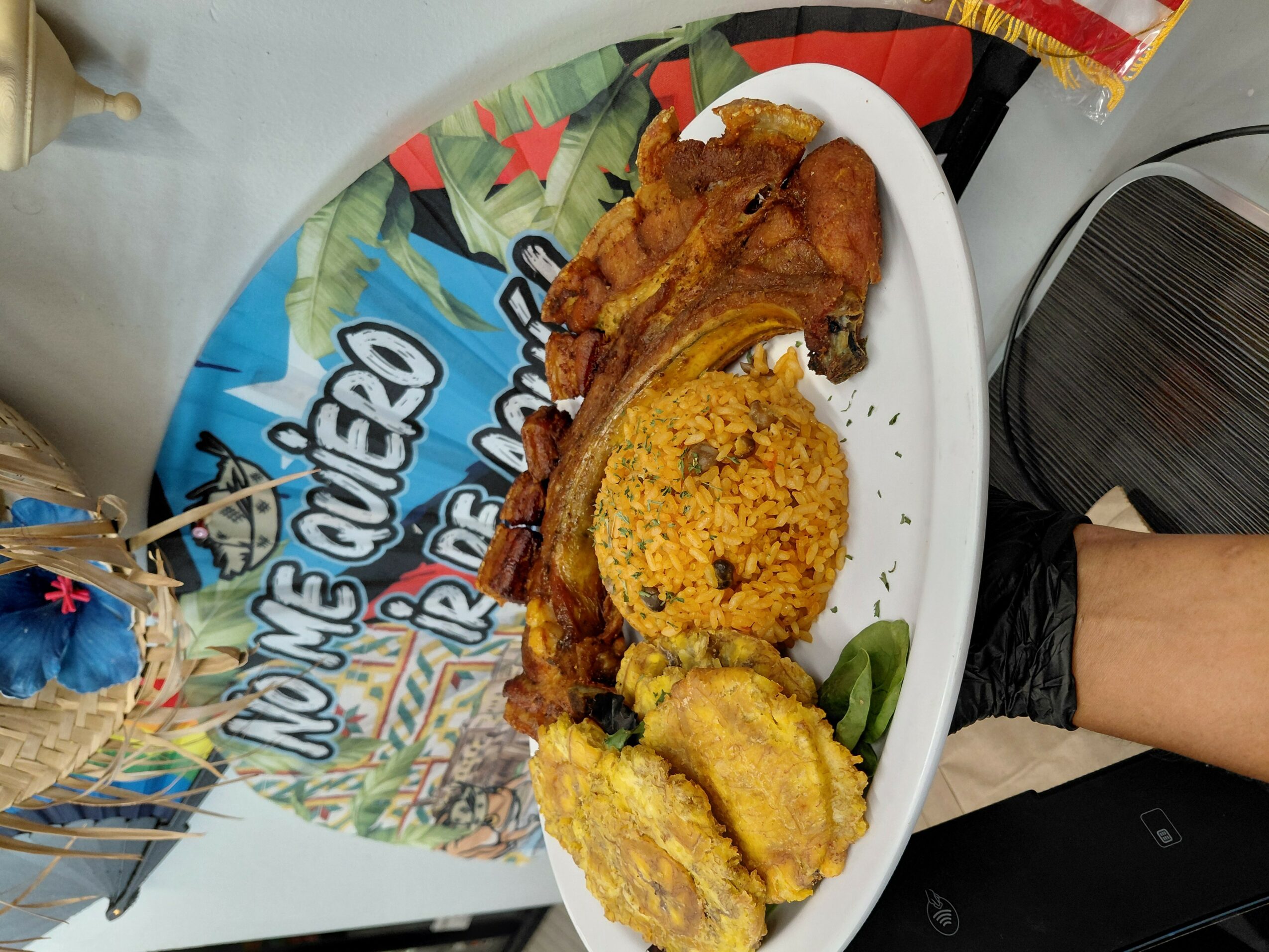 Plate with fried chicken, rice, plantains, and vegetables on a table, with colorful packaging and a person's arm nearby.