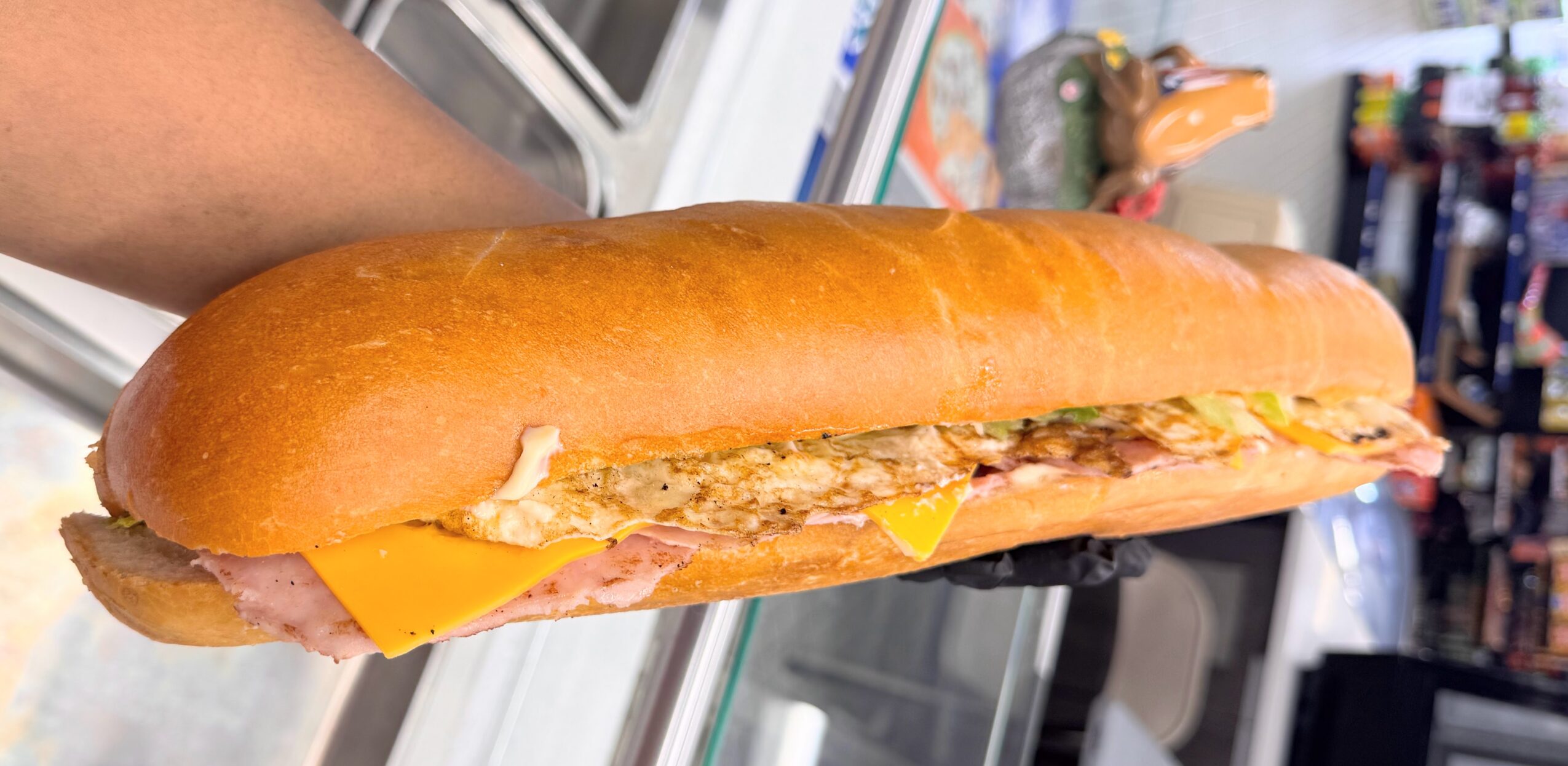 Long sandwich with fried chicken, cheese, and condiments in a bun, held in a hand inside a store.