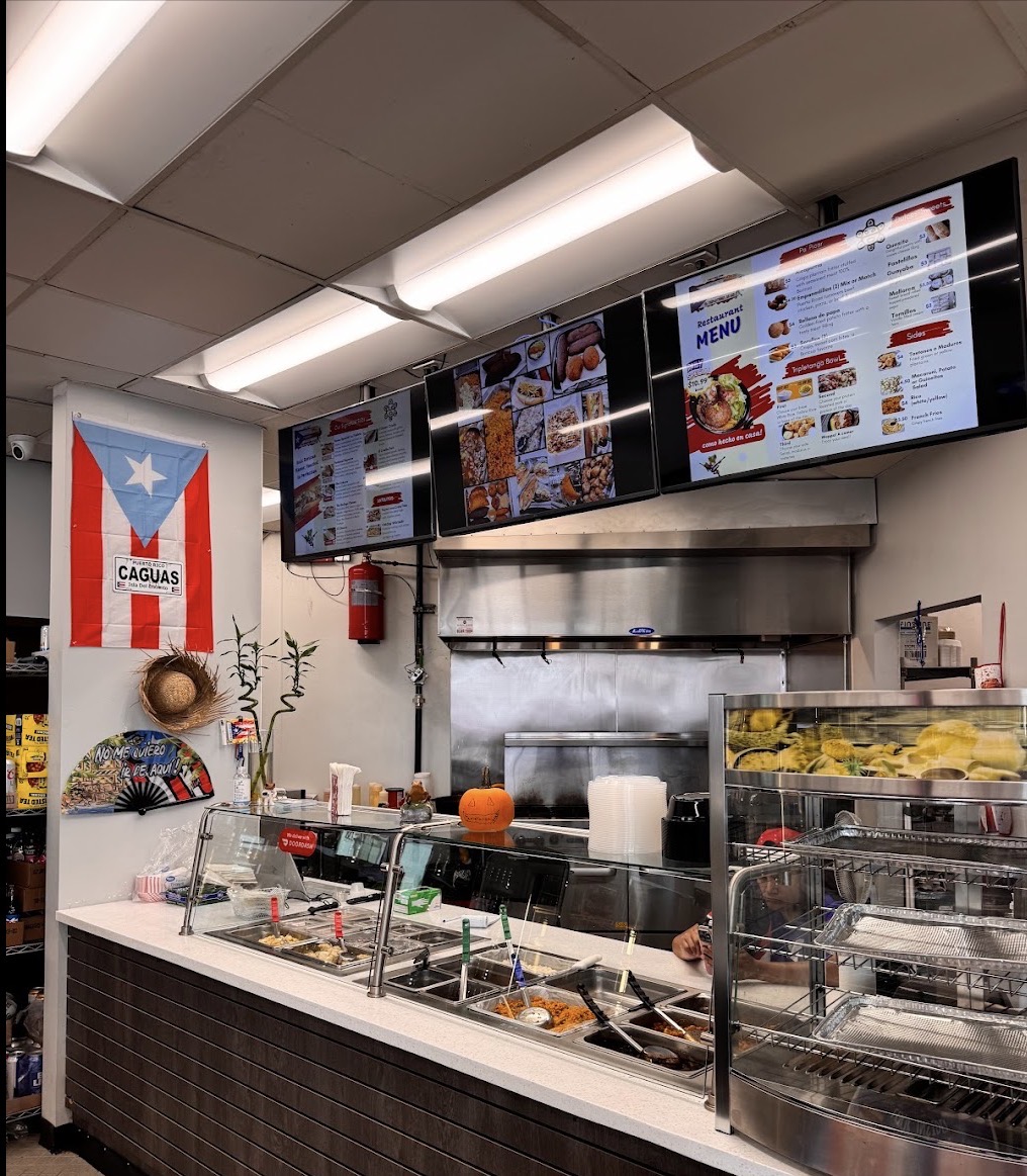View of a restaurant kitchen with digital menu screens, a flag, and various kitchen equipment, seen from the front counter.